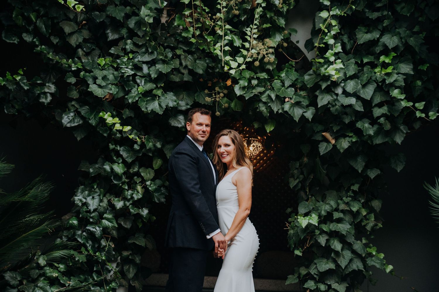 Bride and groom holding hands in front of a lush green ivy wall backdrop on their wedding day.