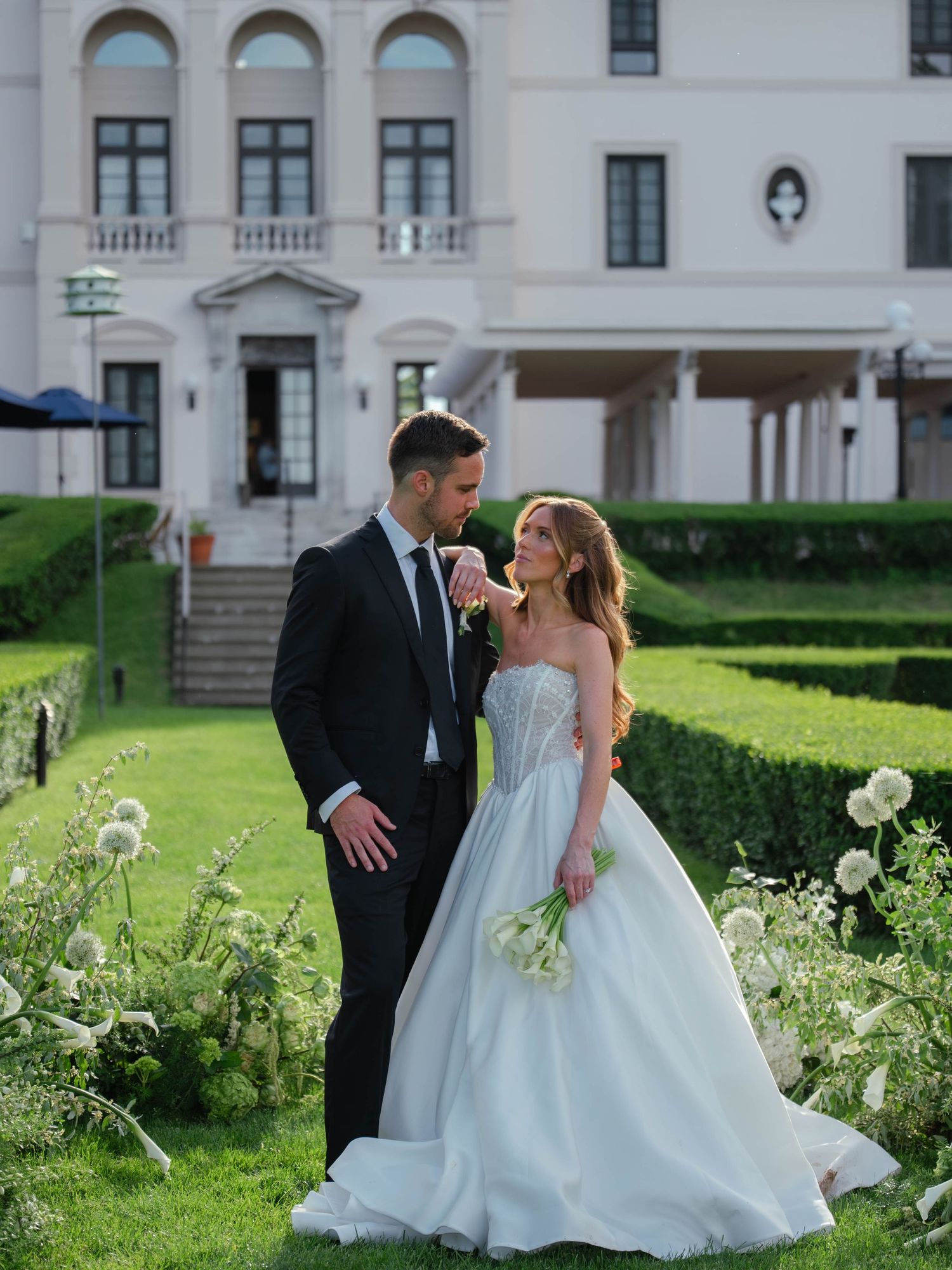 Bride in strapless ball gown and groom in black tuxedo share romantic moment in manicured garden of elegant estate.