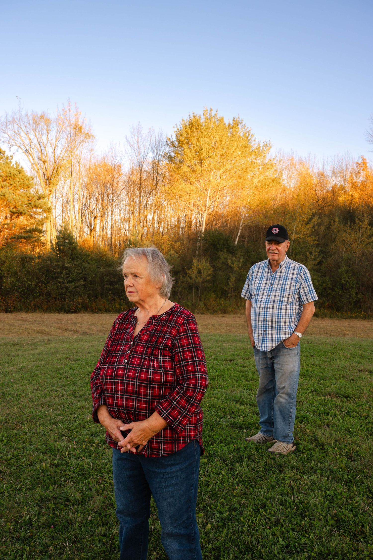 Elderly woman in red plaid shirt stands in field at golden hour with man standing behind her.