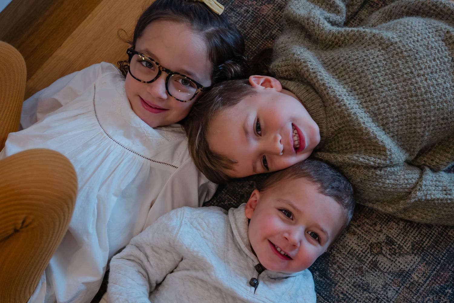 Three young siblings lying together on a cozy blanket, smiling warmly at the camera.