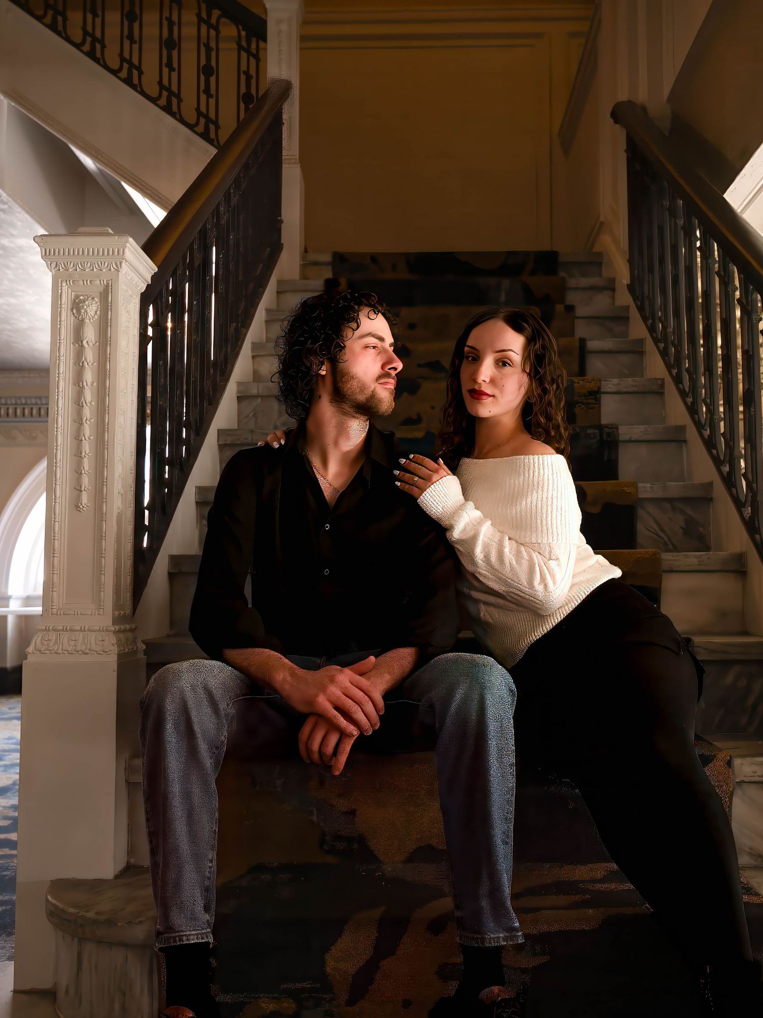 A couple sits on marble stairs in a grand staircase, gazing upward in a romantic pose.