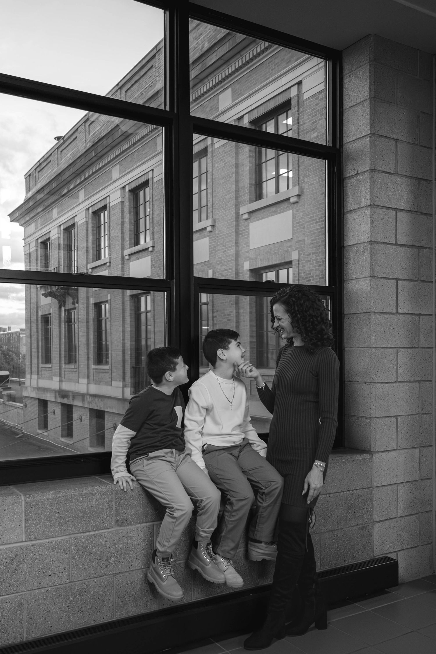 Black and white photo of pregnant mother smiling at two boys on a window ledge indoors.