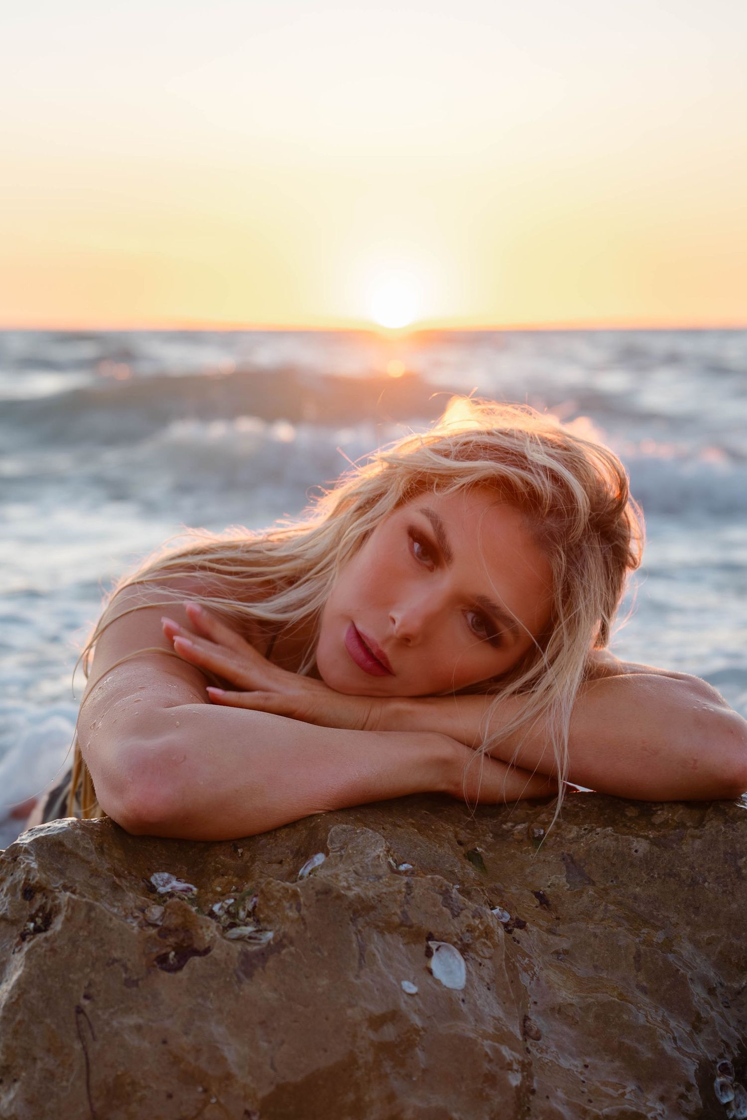 Blonde woman rests chin on arms atop seaside rock during warm ocean sunset.