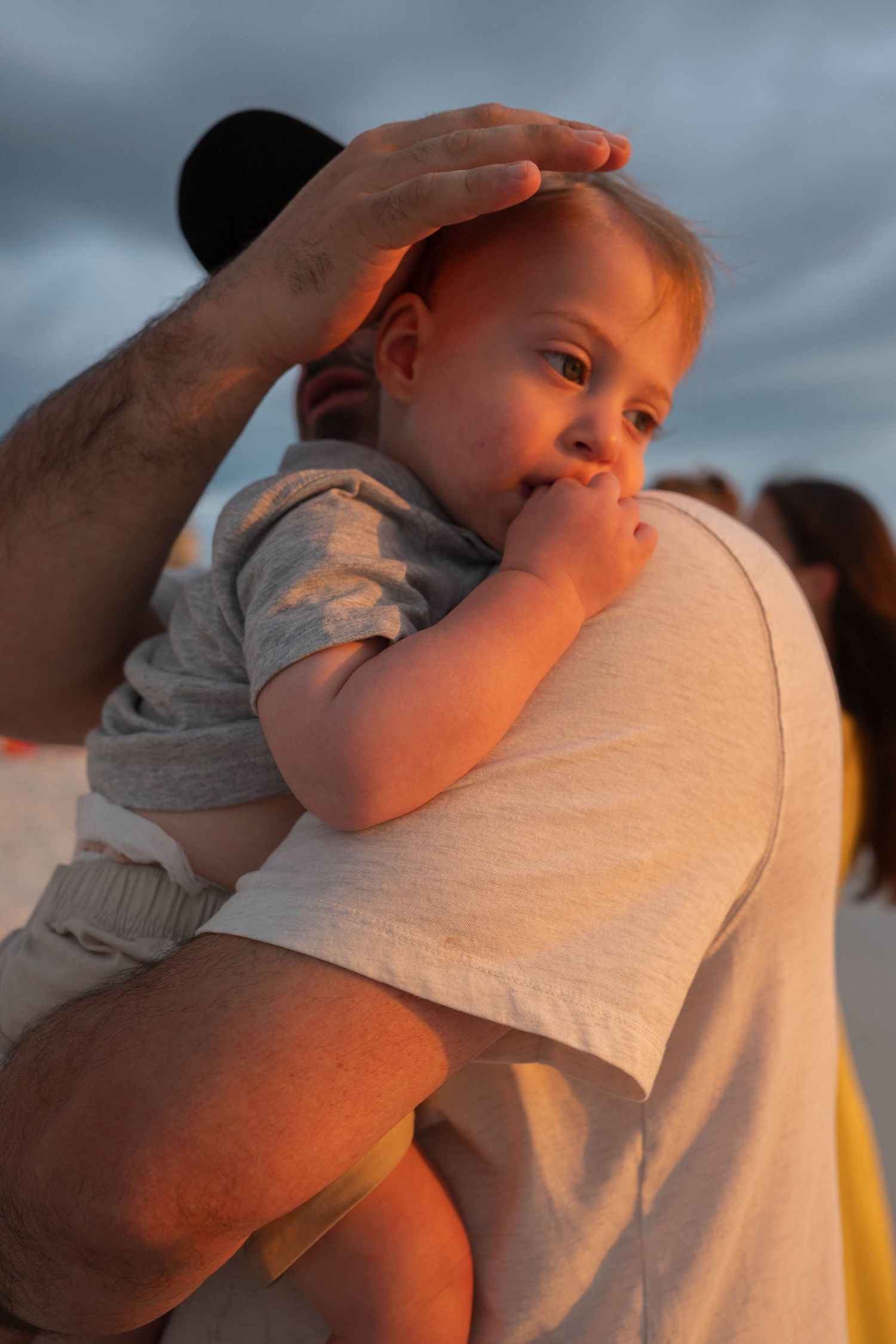 A man holds a baby in golden sunset light outdoors, gently placing his hand on the infant's head.