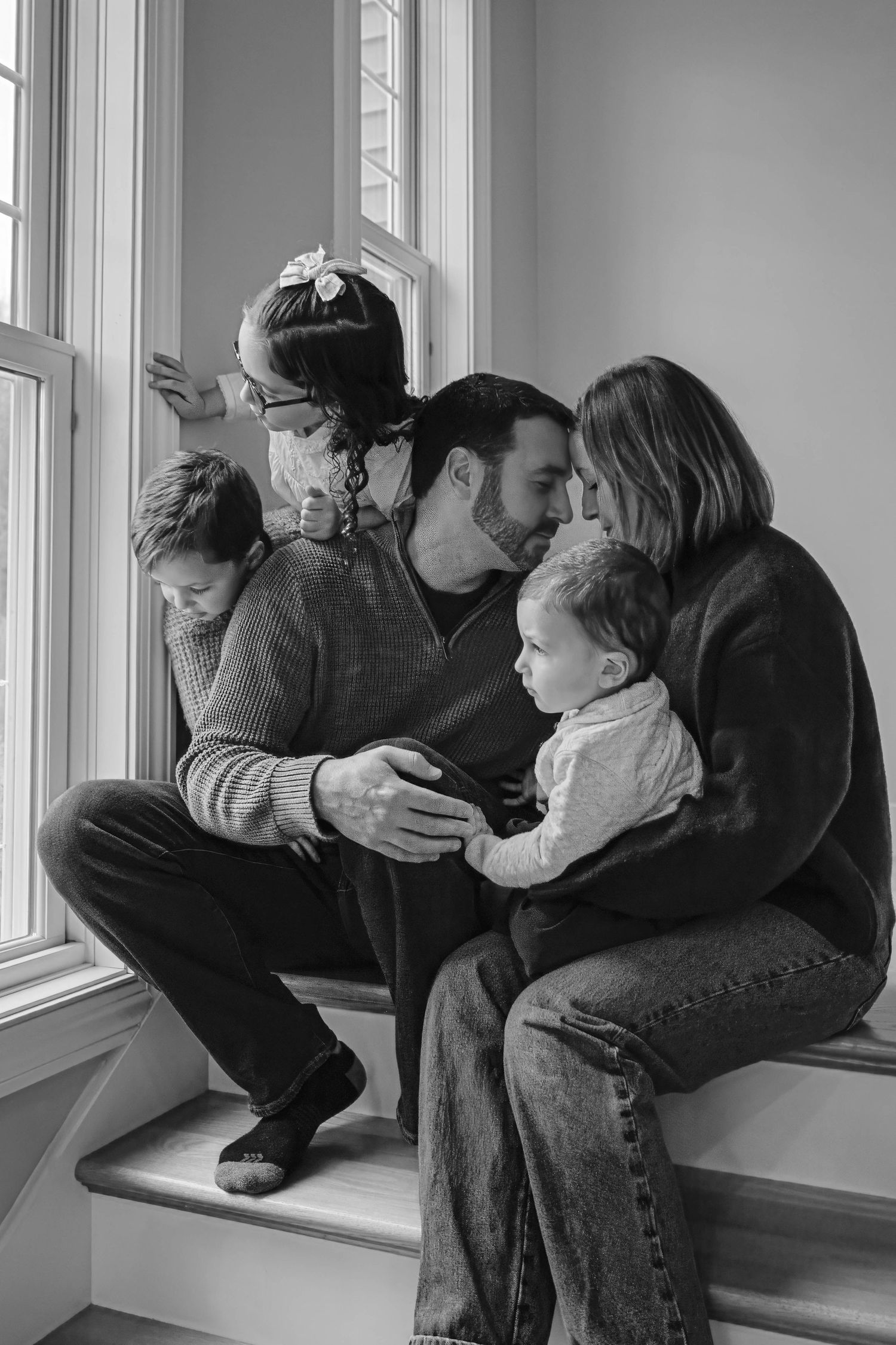 Black and white family photo of parents with three young children sitting by a window on indoor steps.