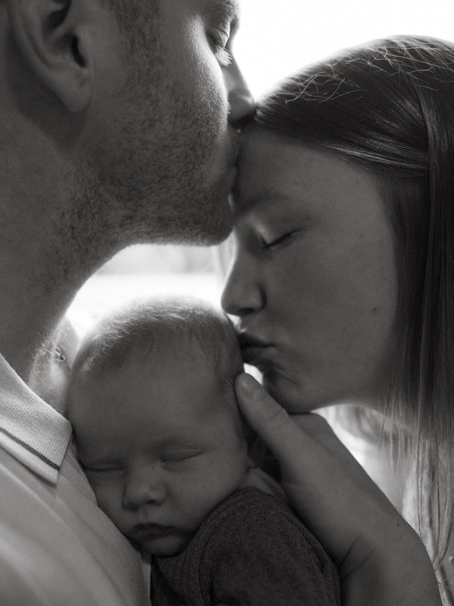 Black and white photo of two parents kissing over their sleeping newborn baby, a tender family portrait.