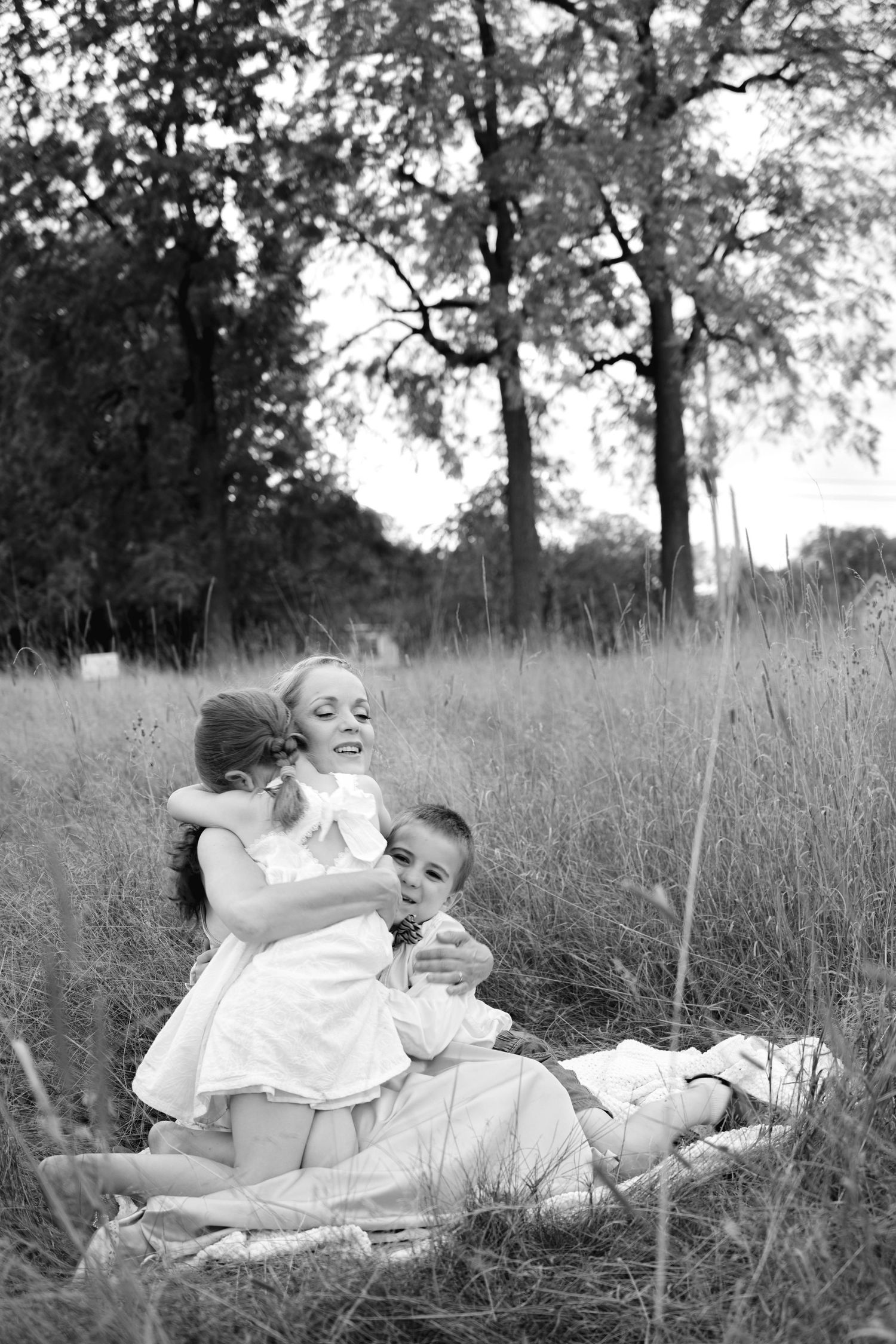 Black and white photo of a mother and two children hugging in a grassy field with trees in background.