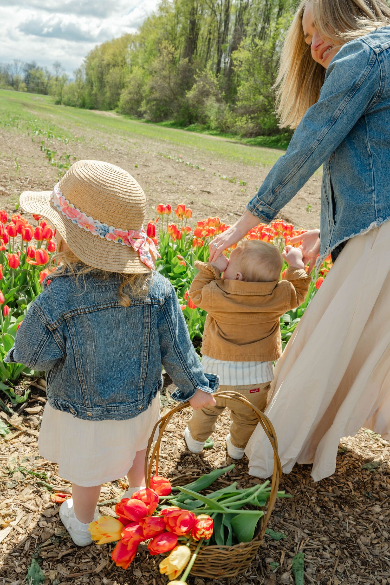 Mother guiding two young children with tulip basket along colorful spring flower field path.