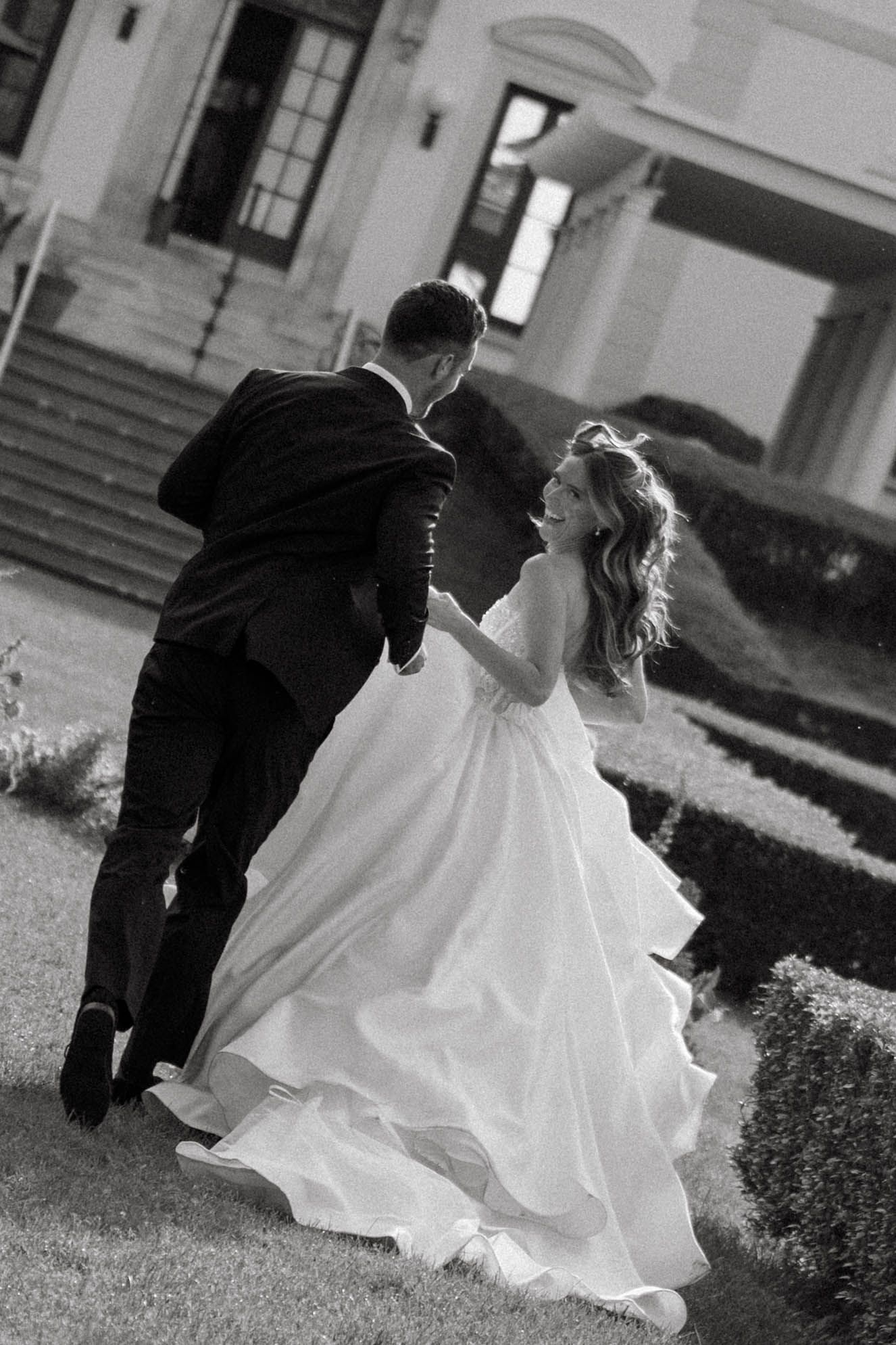 Black and white photo of laughing bride in ball gown running with groom in formal suit outdoors.