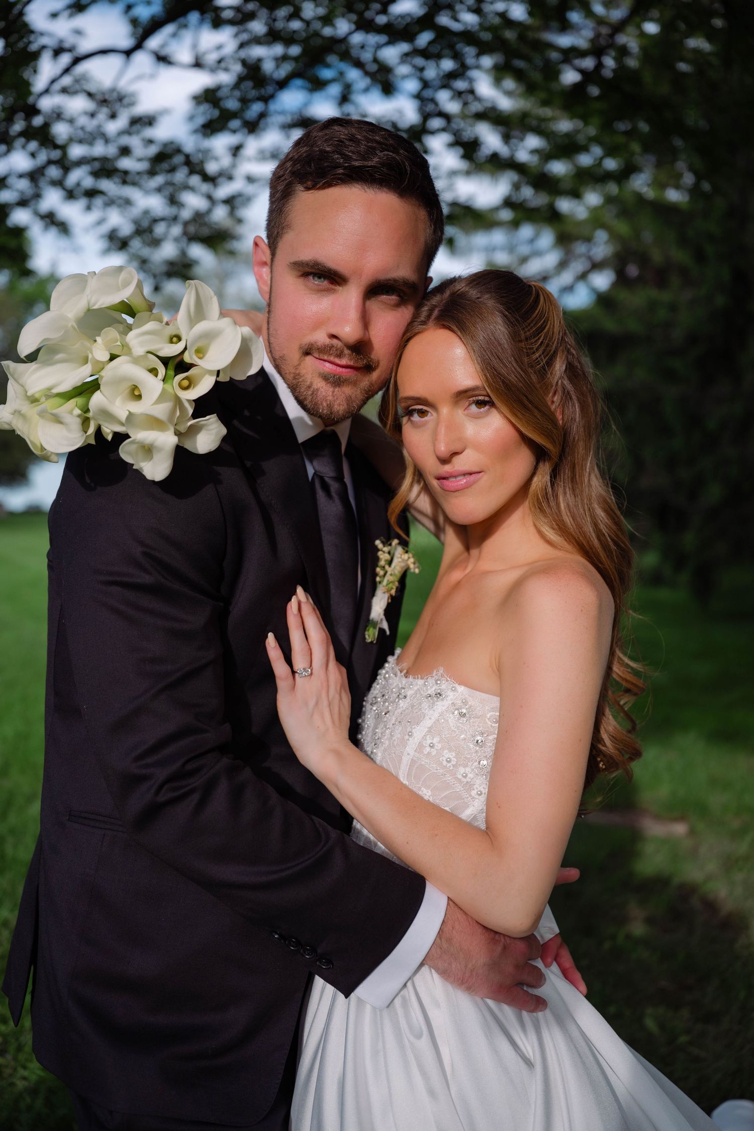 Bride and groom posing together holding white calla lily bouquet in a sunlit outdoor garden.