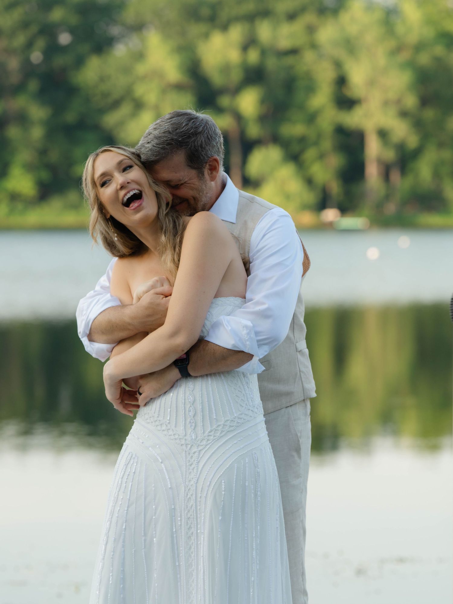 Happy couple embracing and laughing by a lakeside, woman in white wedding dress, man in light suit, lush green trees background.