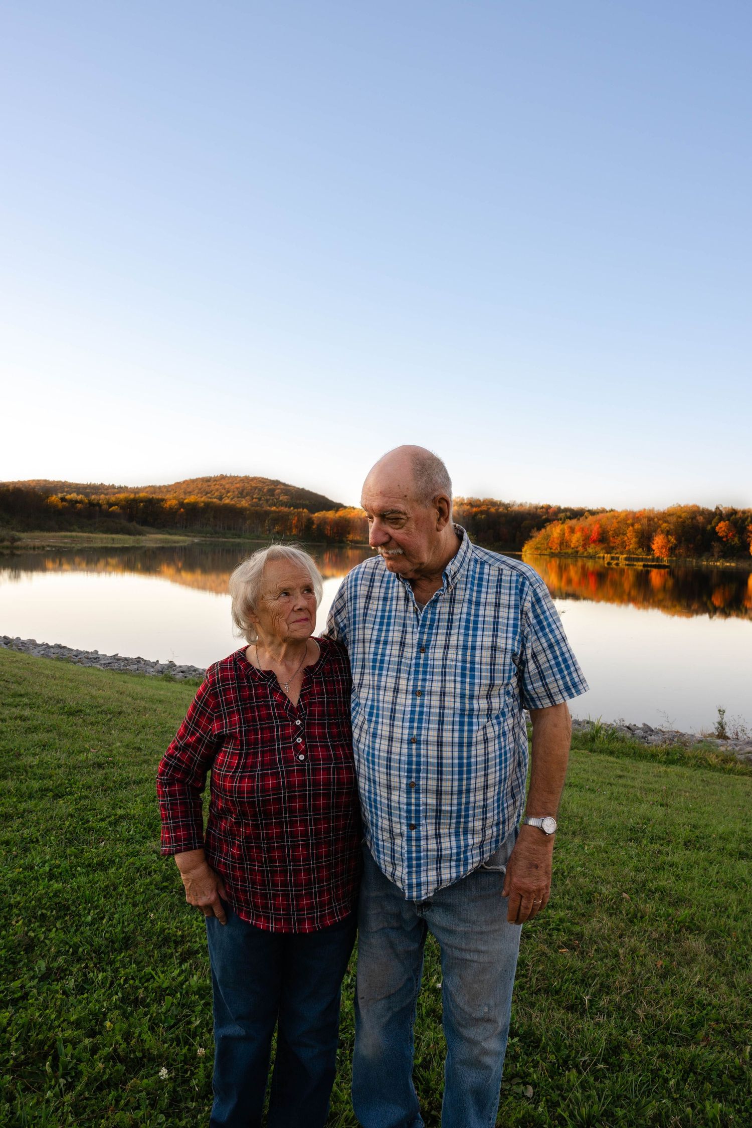 Elderly couple standing together by a reflective lake with autumn foliage in background at sunset.