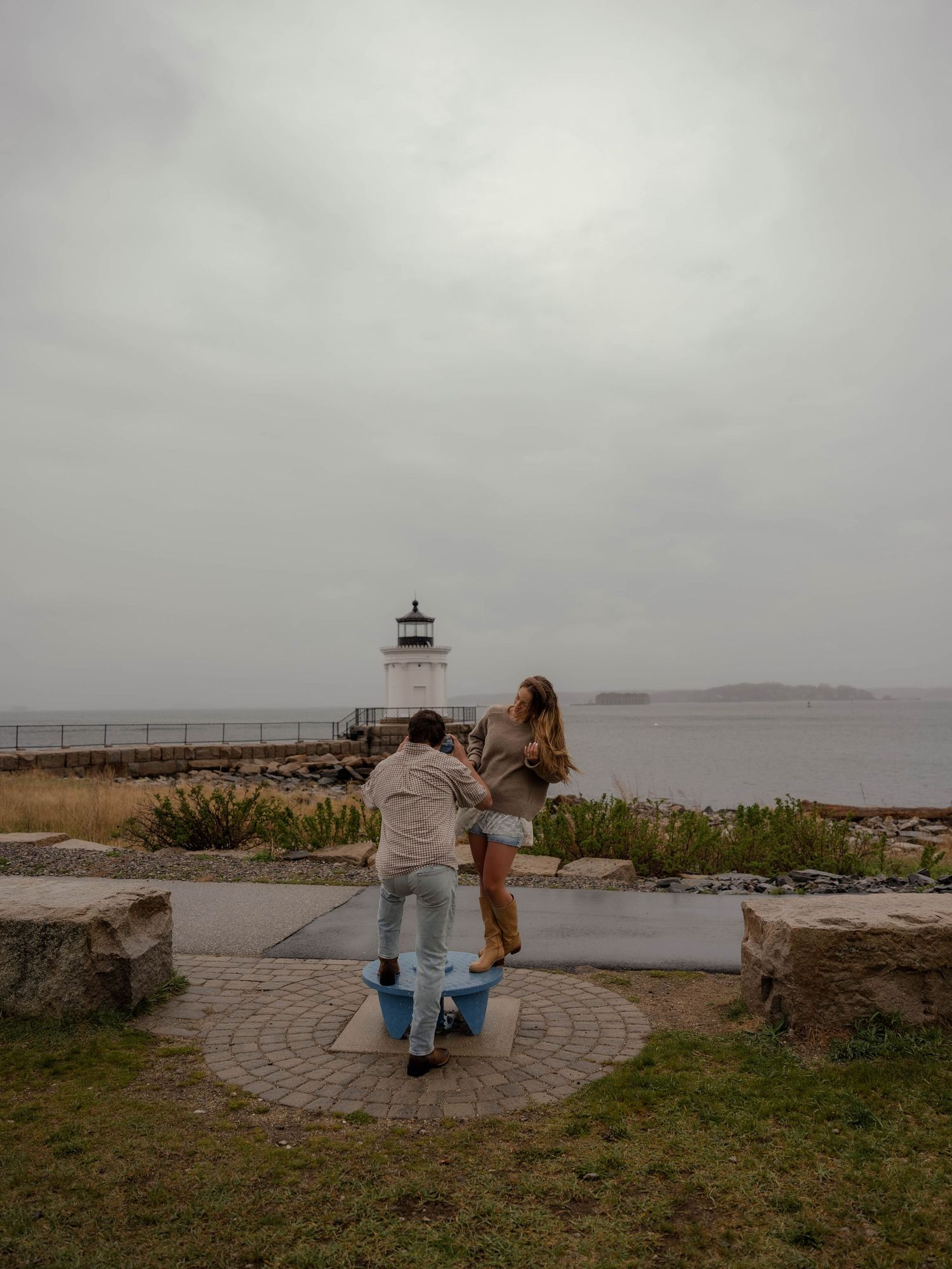 Couple dances playfully on coastal plaza with white lighthouse and gray ocean in background.
