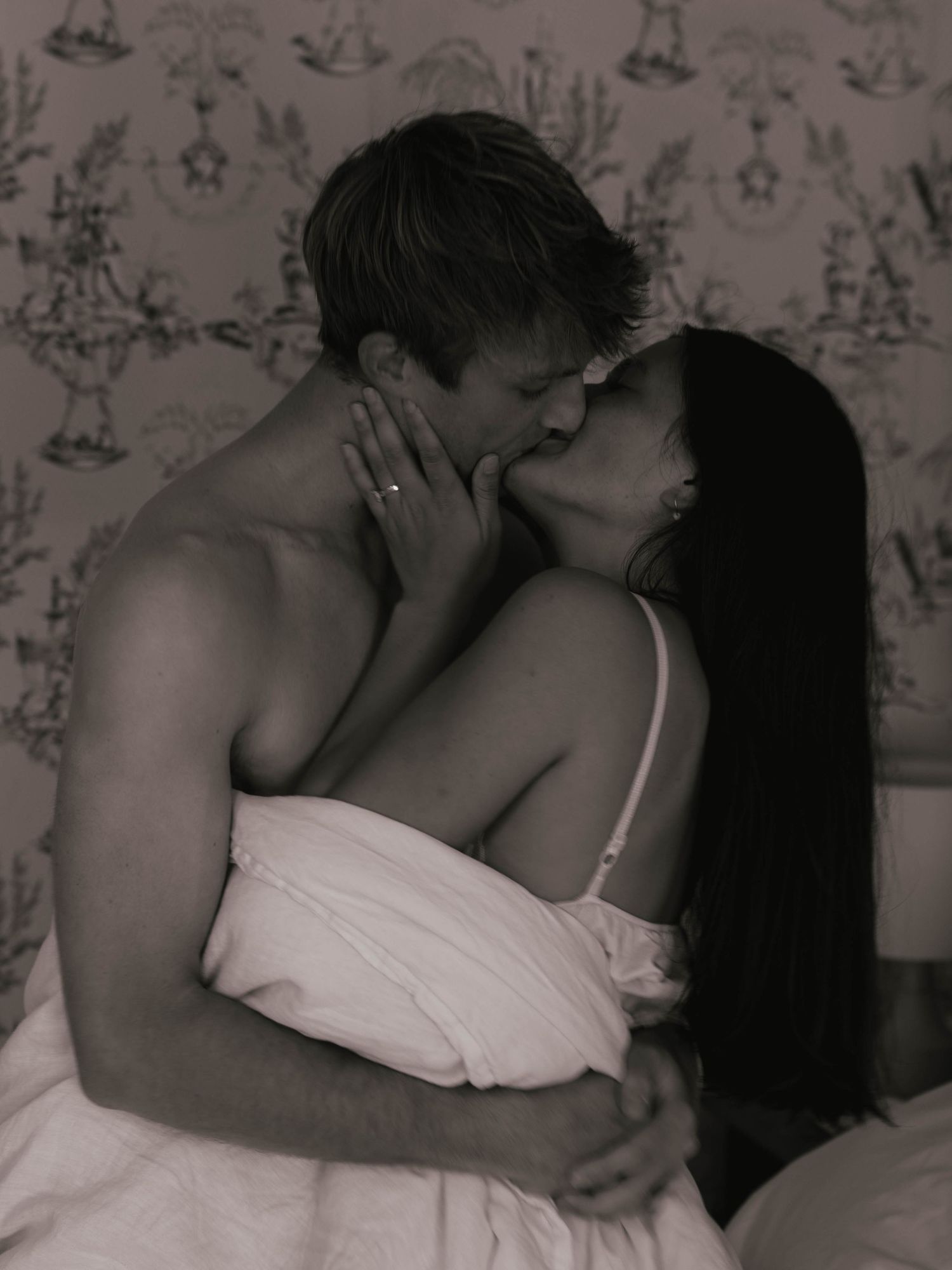 Black and white photo of passionate couple kissing in bed with decorative wallpaper background.