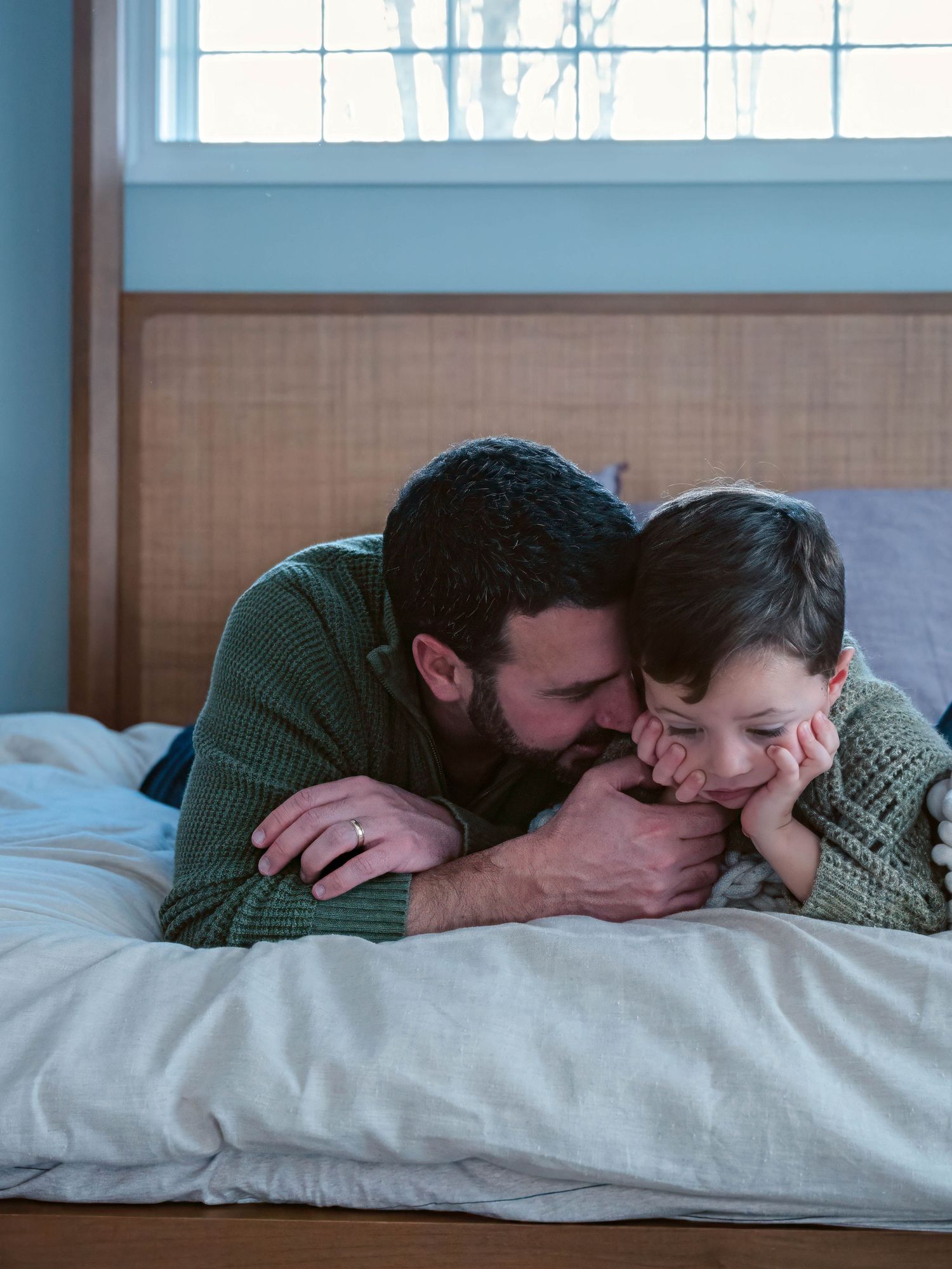 A loving father in a green sweater cuddles his young son on a bed by a window.
