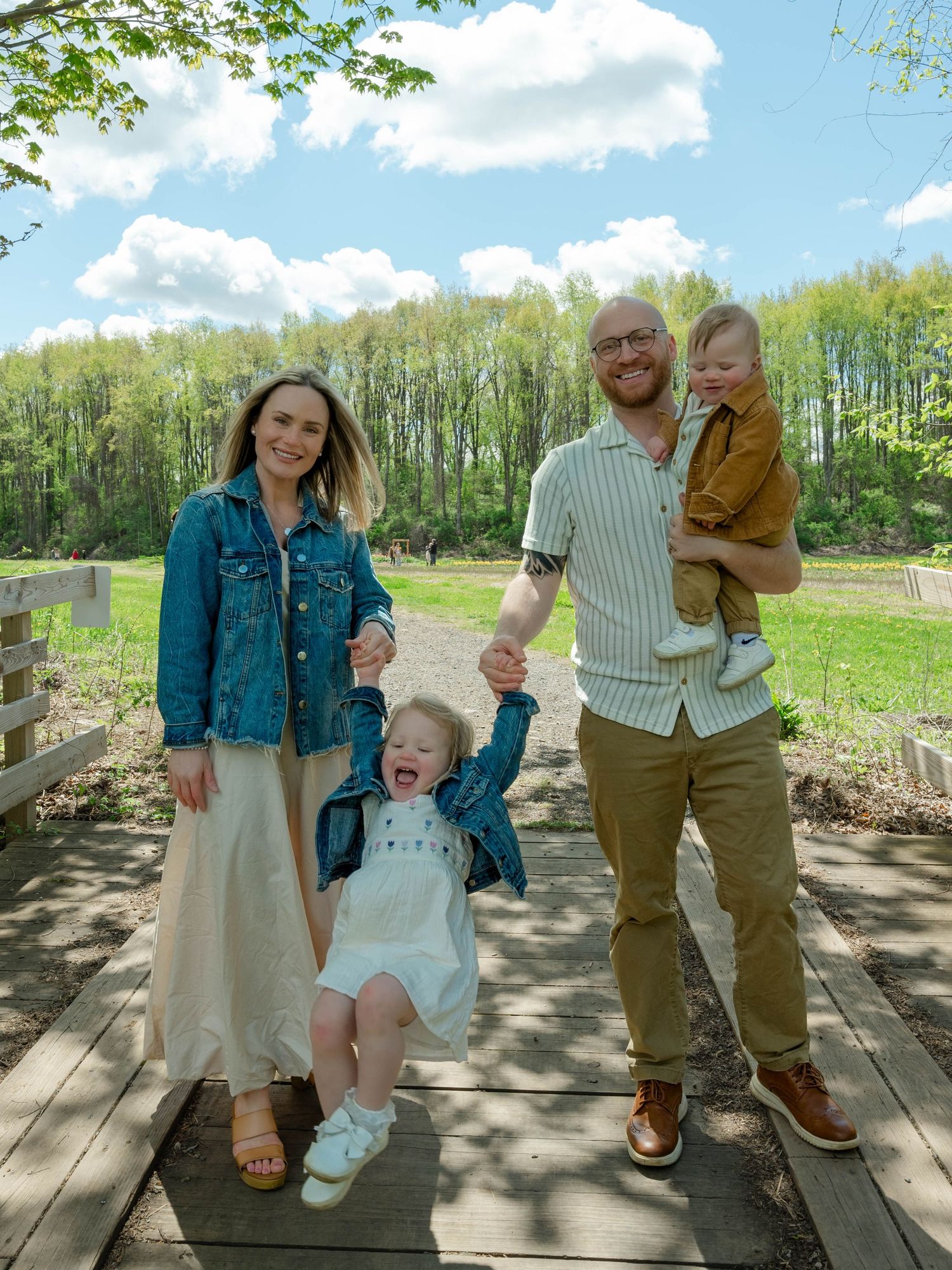 Happy family of four laughing and swinging toddler on sunny wooden bridge surrounded by lush green trees.