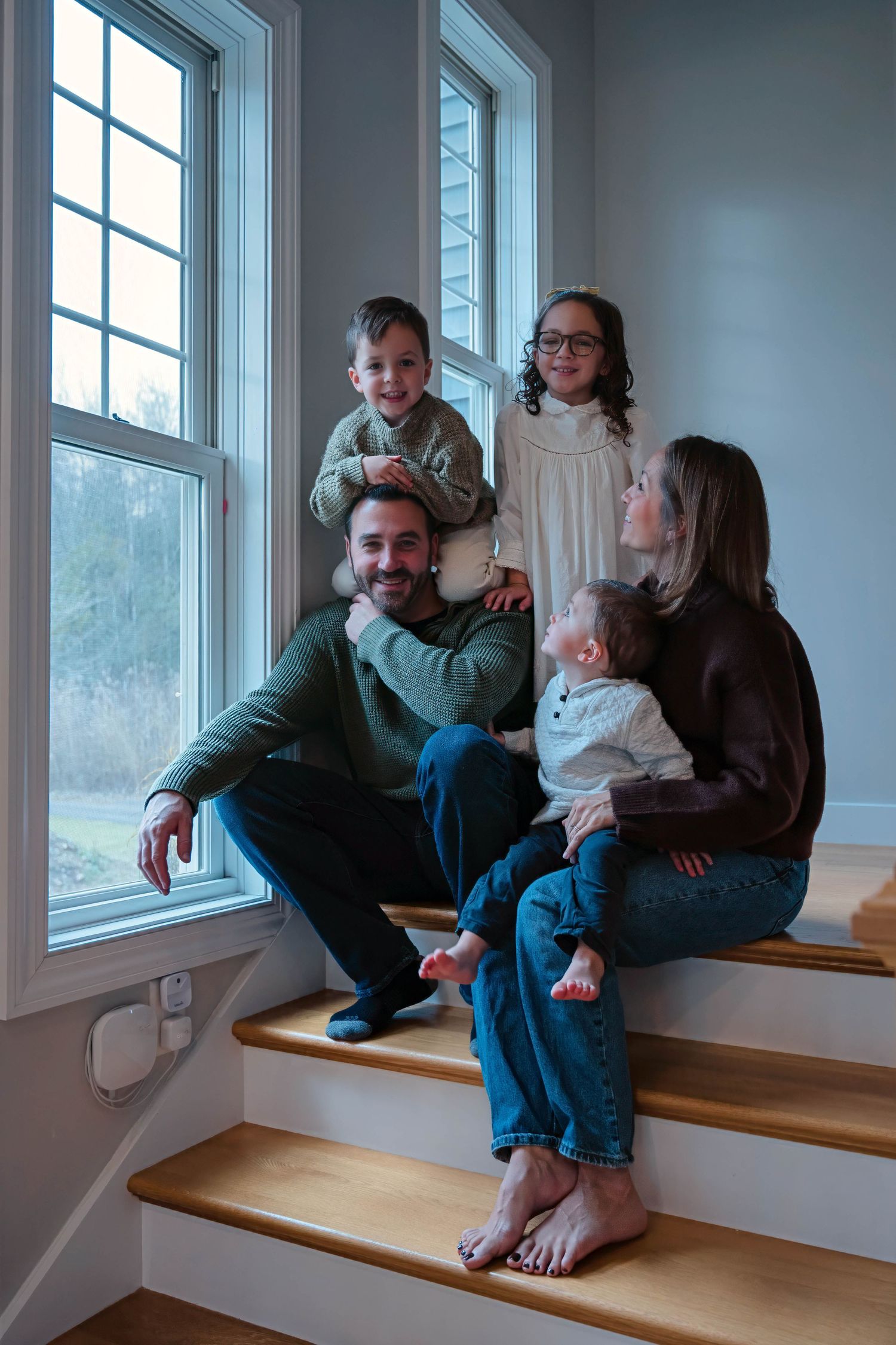 Happy family of five poses on wooden staircase steps by large windows in bright home interior.