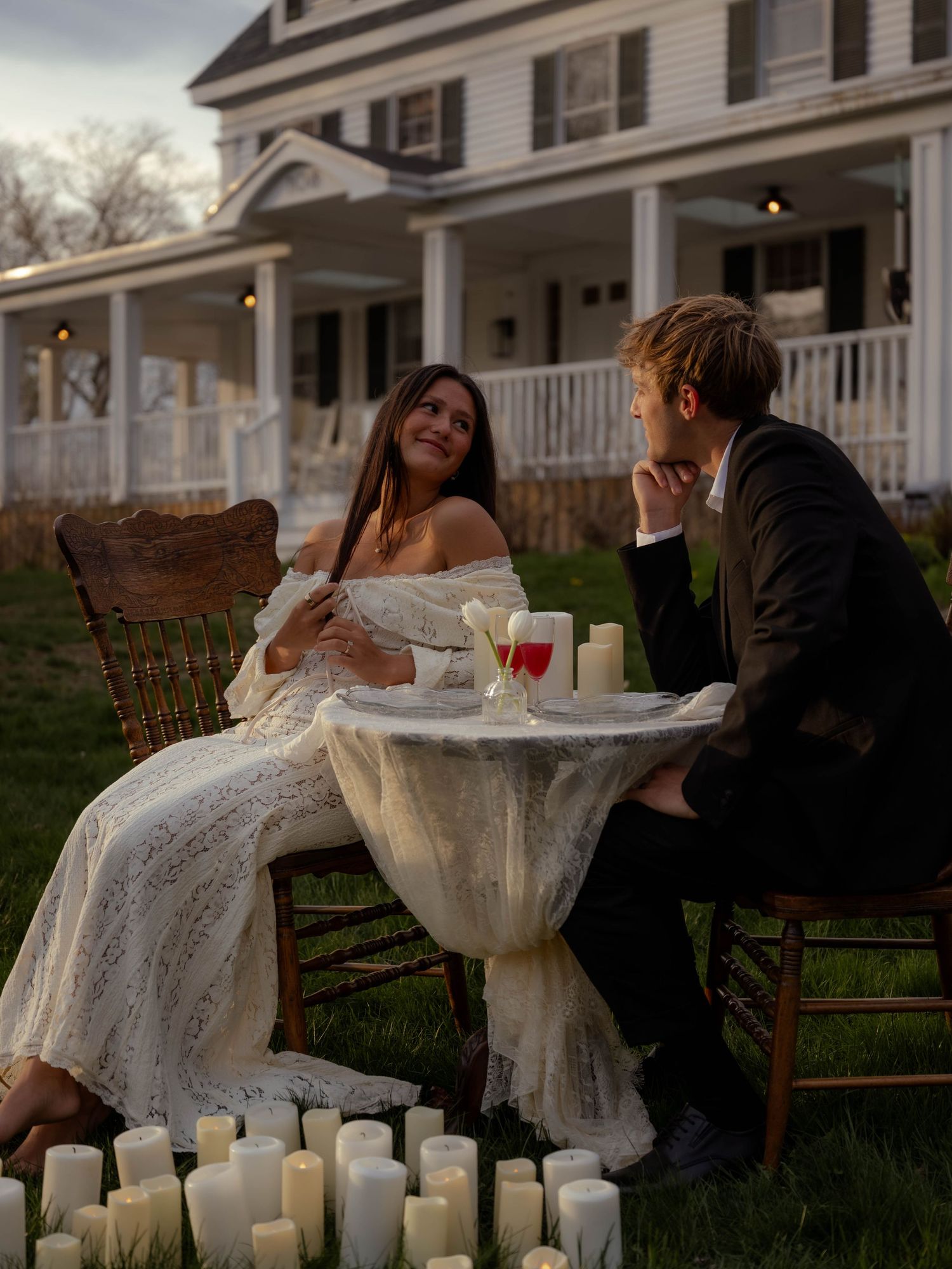 Bride and groom share candlelit outdoor evening table setting surrounded by white pillar candles at rustic farmhouse.