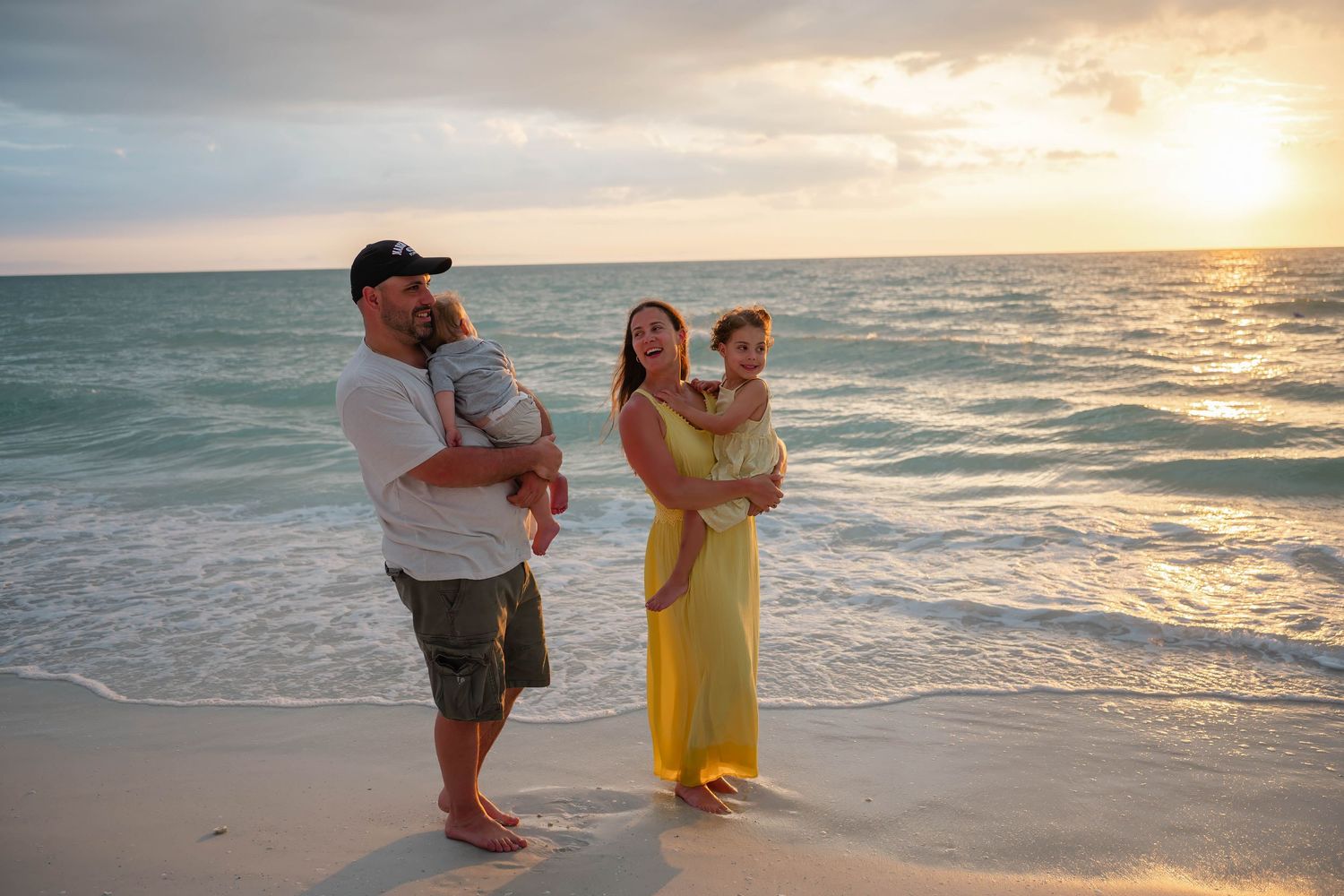 Family of four poses on a sandy beach at sunset, parents holding toddlers in yellow dress and casual wear.