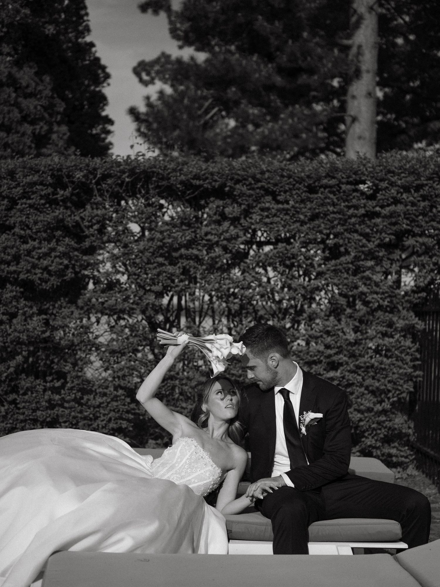 Black and white romantic photo of bride raising bouquet while groom leans in close outdoors.
