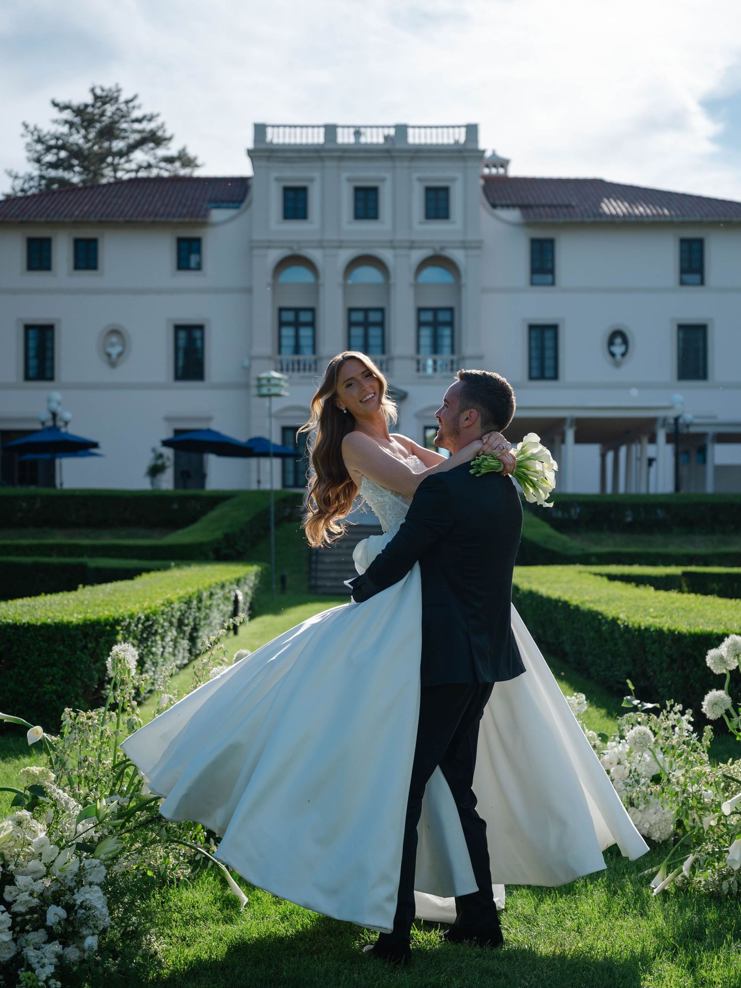 Groom lifts bride in white ballgown in manicured garden in front of grand estate wedding venue.