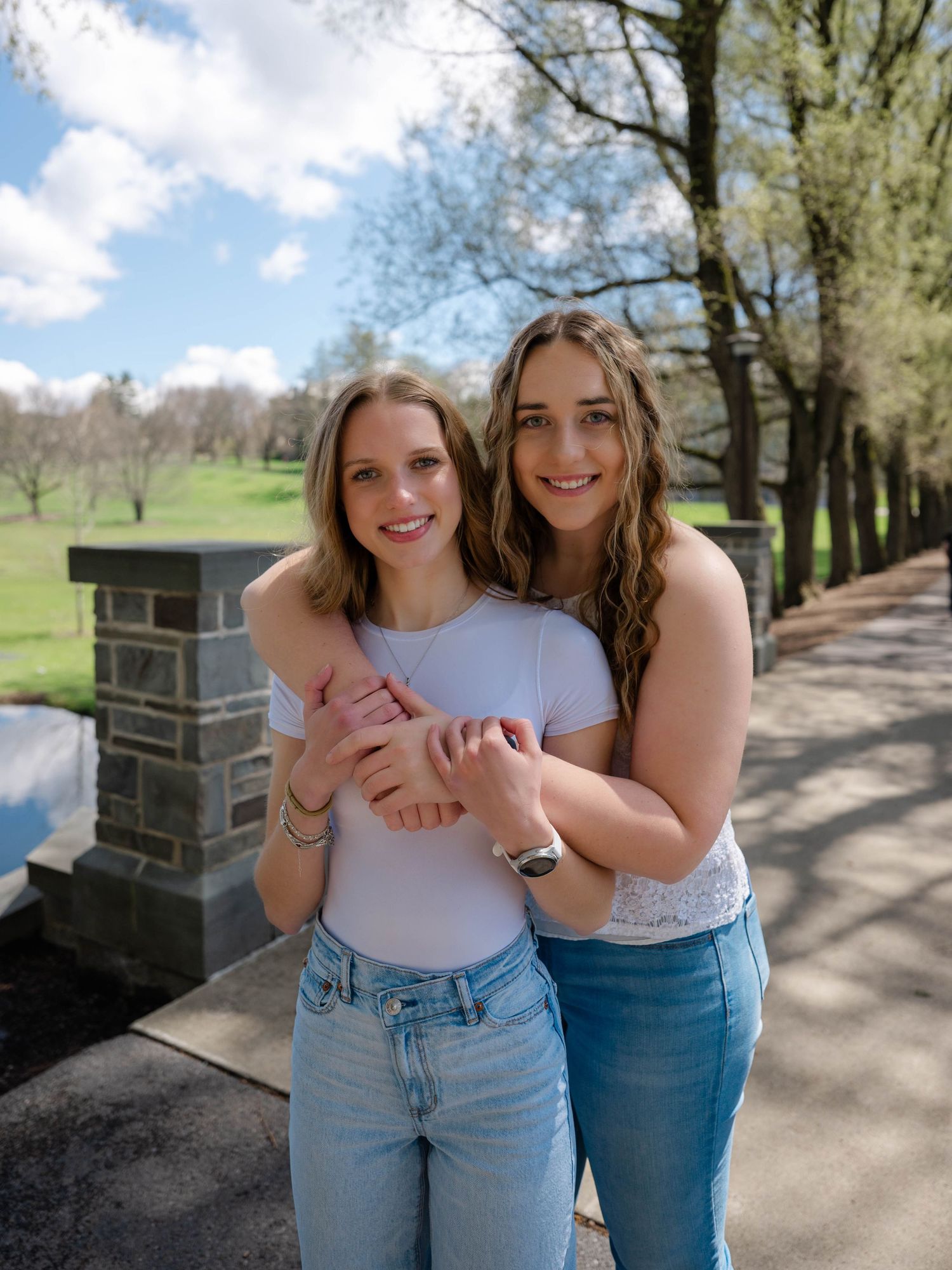 Two young women smile and embrace on a sunny park pathway with blooming spring trees behind them.