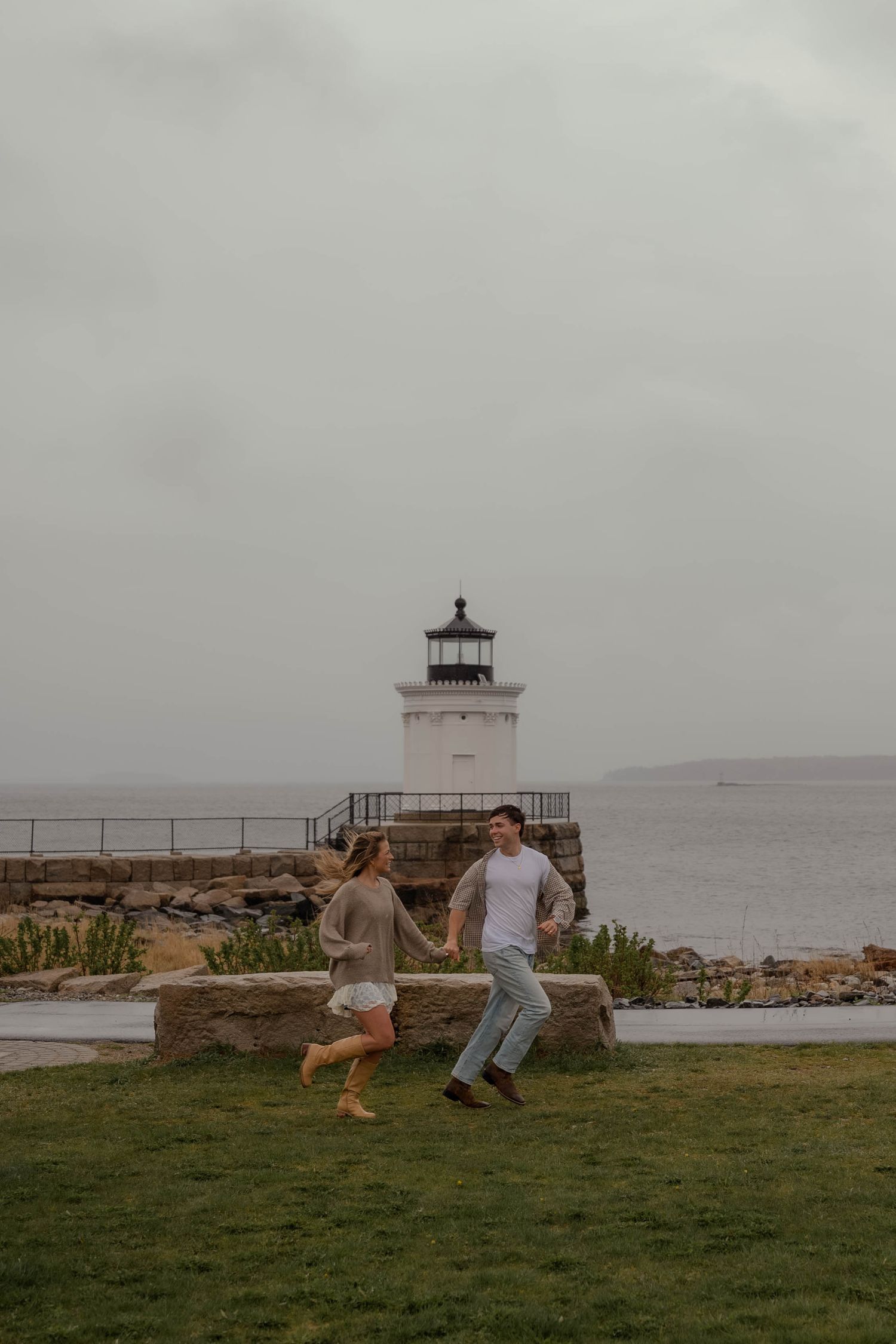 Joyful couple running hand in hand on grass with white lighthouse behind them.