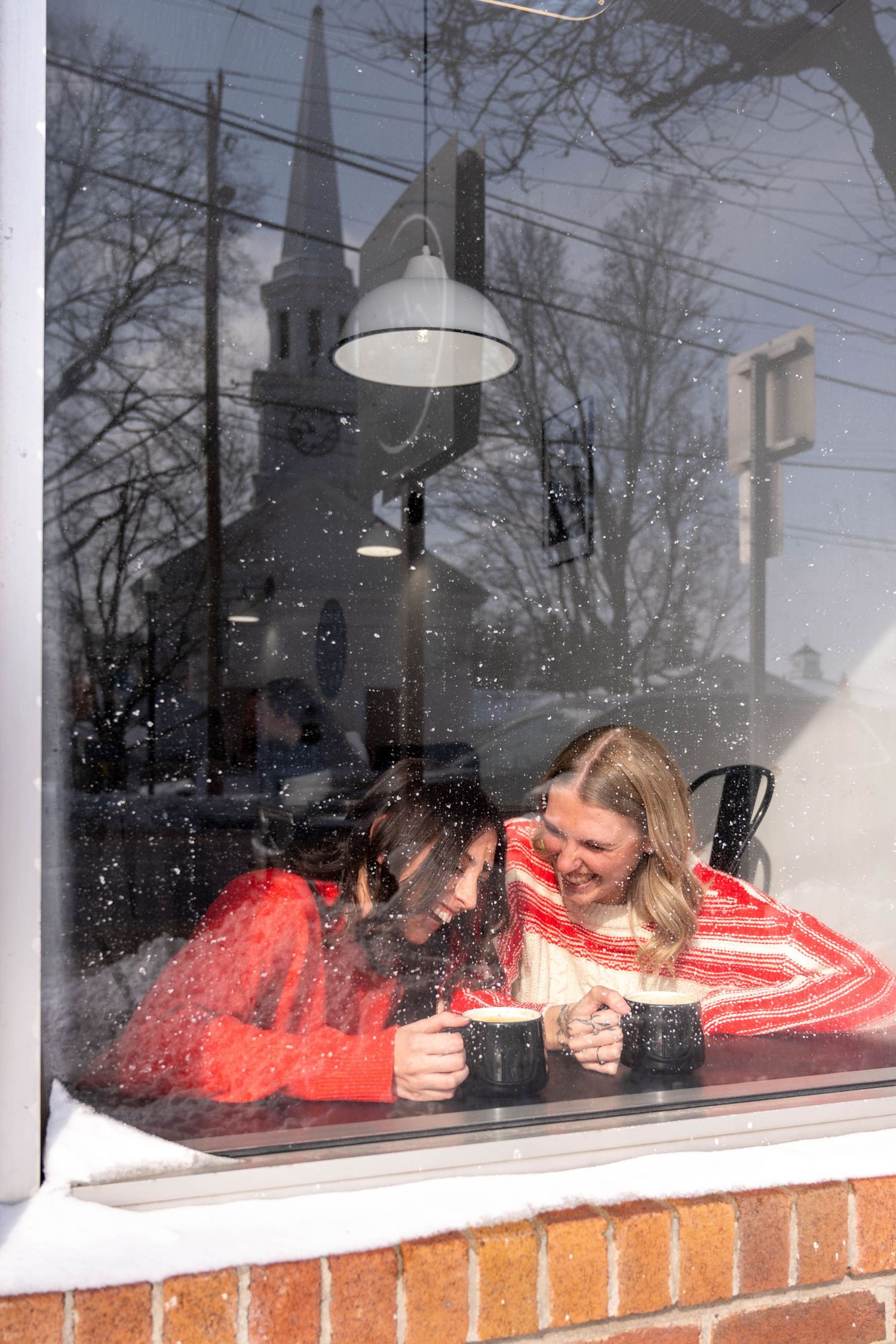 Two women laughing together inside a cafe, viewed through a snowy winter window.