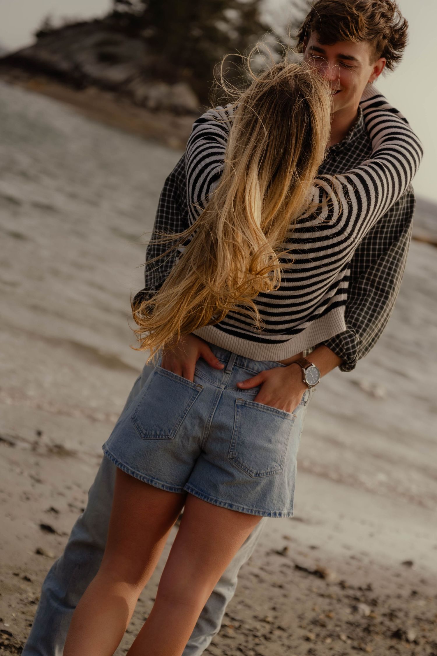 Couple embracing warmly on a sandy beach with rocky shoreline in the background.