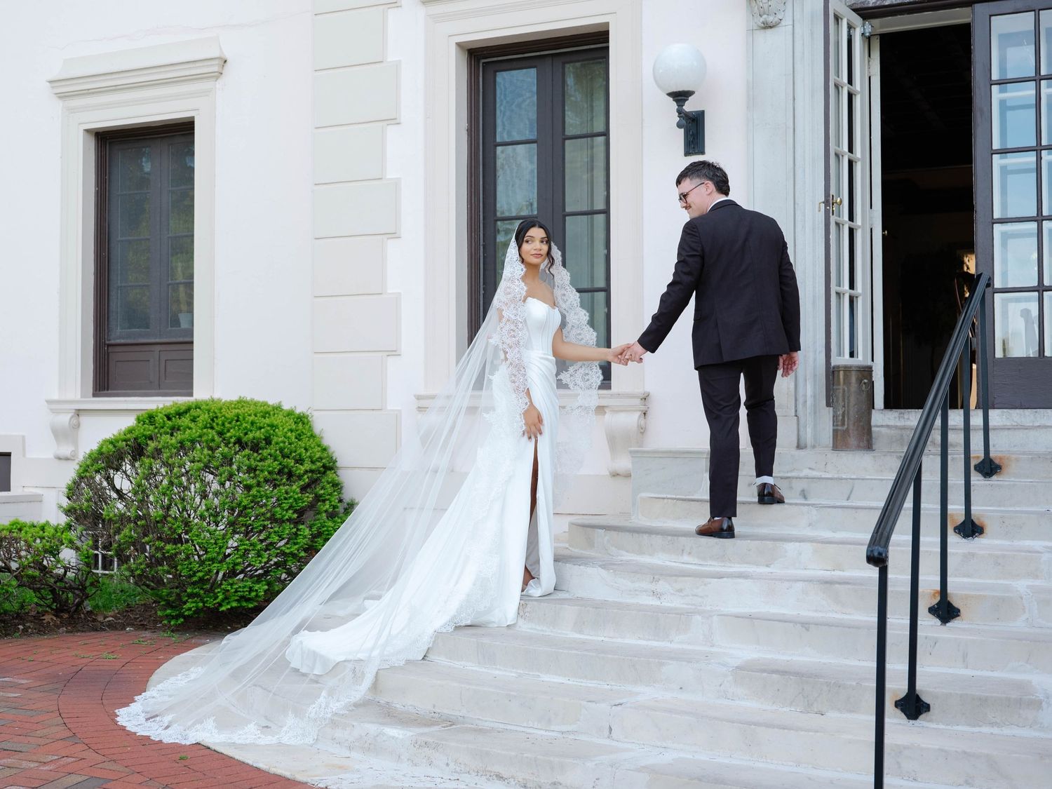 Bride in a white gown with cathedral veil holds grooms hand on steps of elegant white venue.