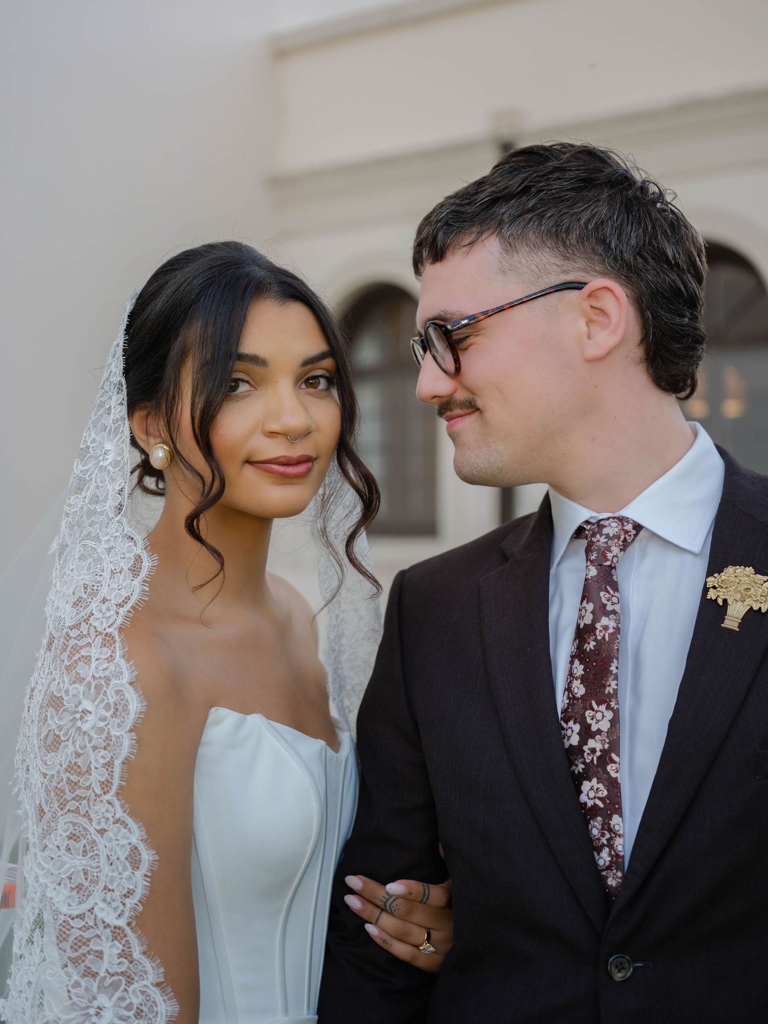 Radiant bride in strapless gown with lace veil smiles beside groom in dark suit and floral tie.