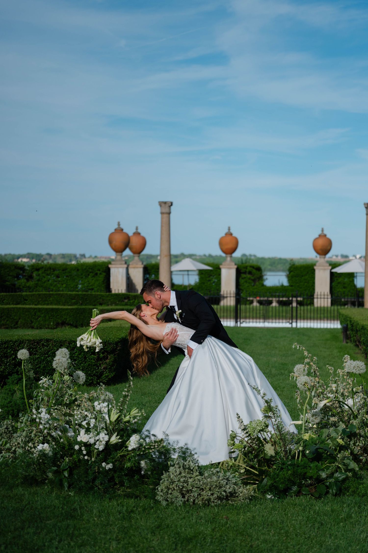 Groom dipping bride in white ballgown for a kiss among white flowers in sculpted garden landscape.
