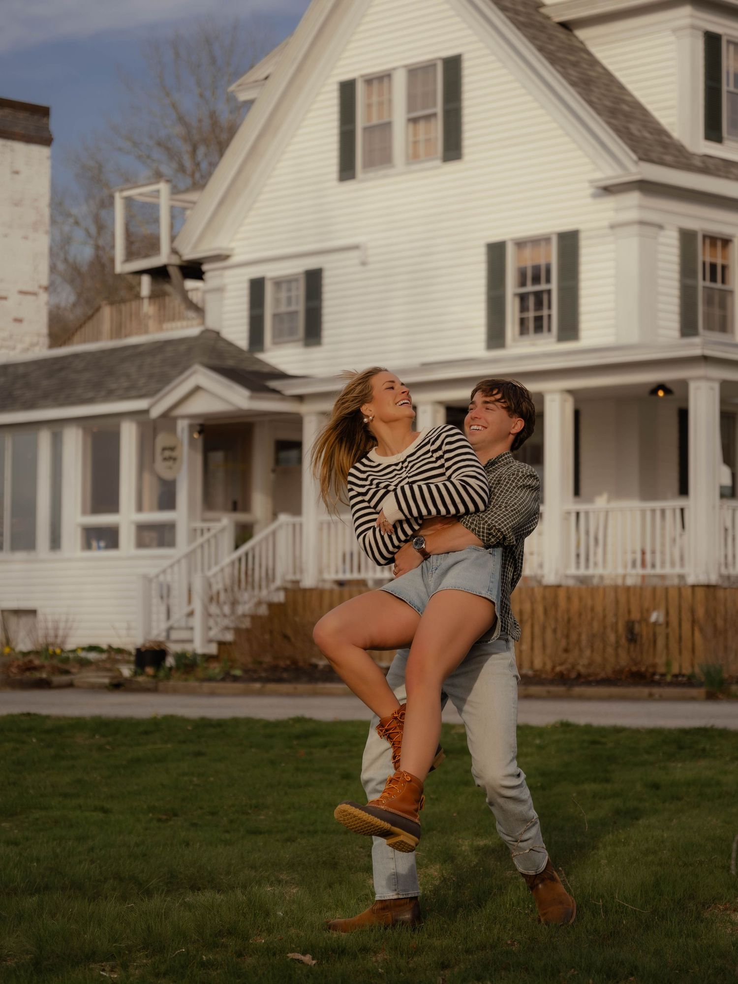 Joyful couple embracing and spinning in front of a classic white New England colonial home.