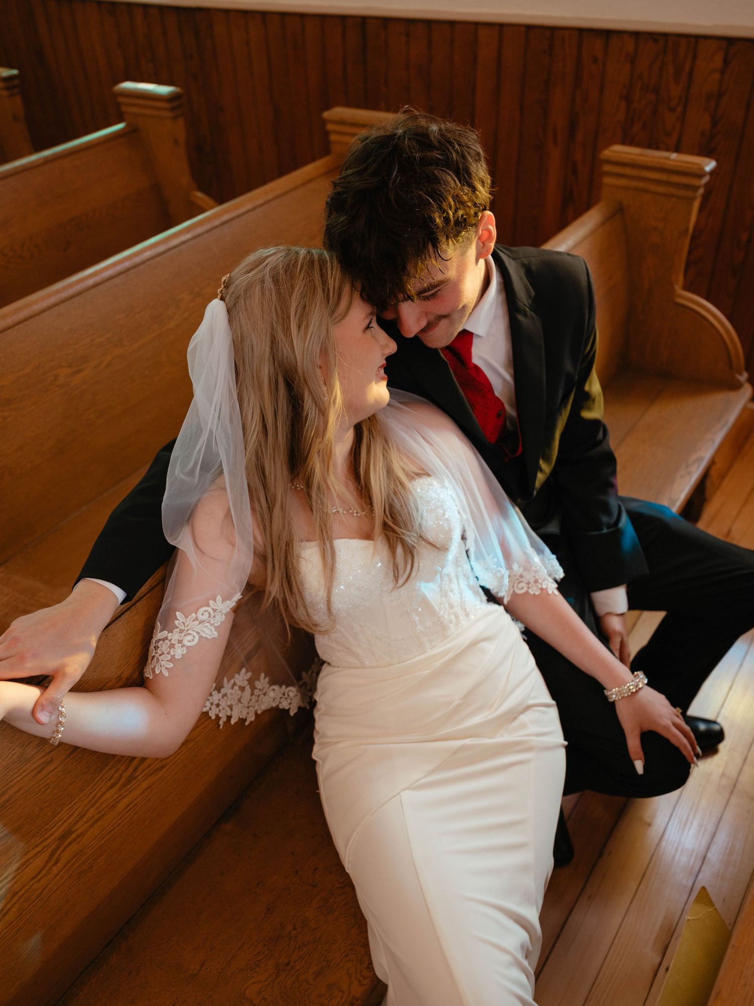 Bride and groom share an intimate moment seated on wooden church pews, bride in white gown with veil, groom in black suit with red tie.