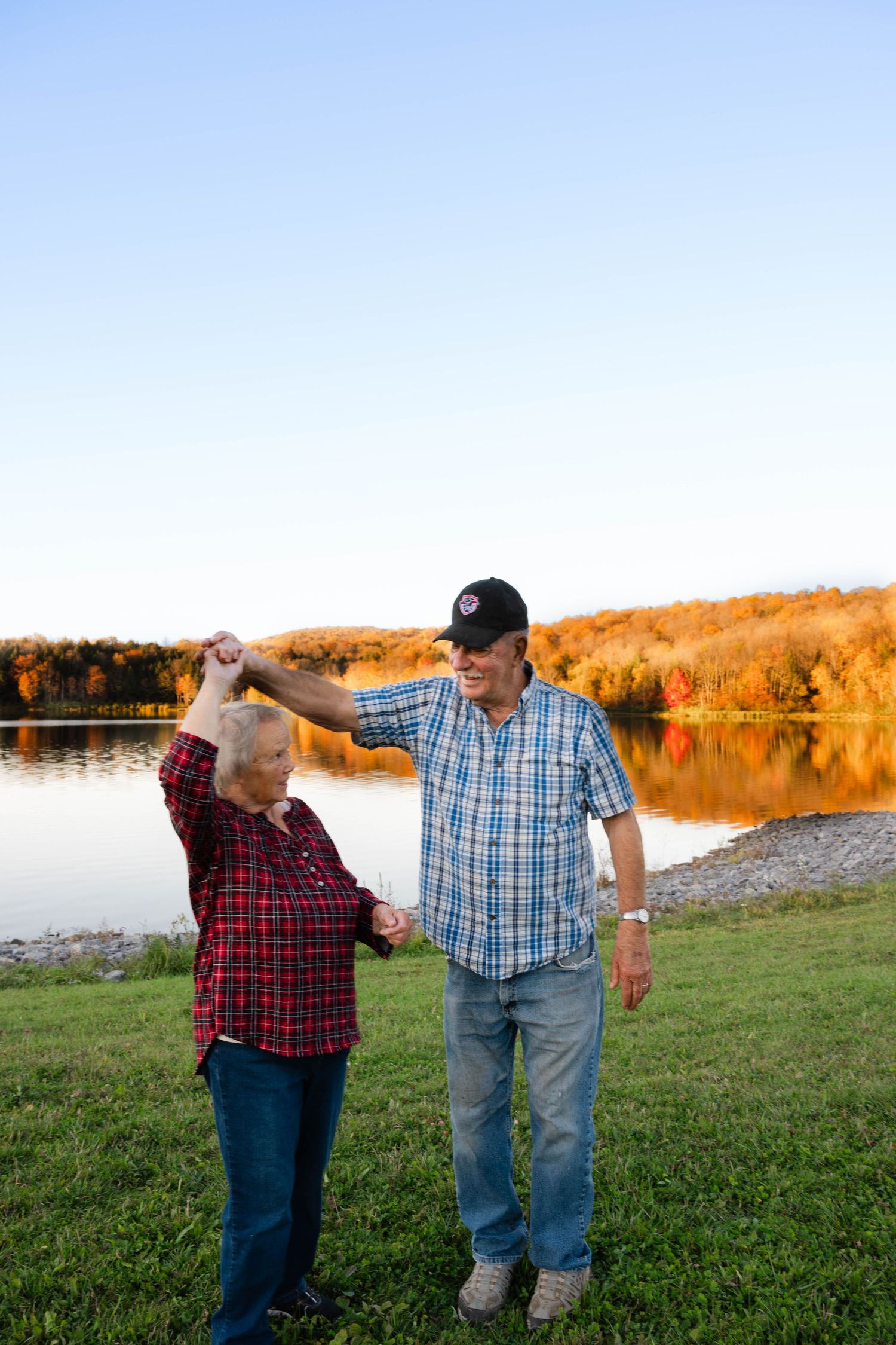 Elderly man twirling woman by hand near scenic autumn lake at golden hour.Central New York