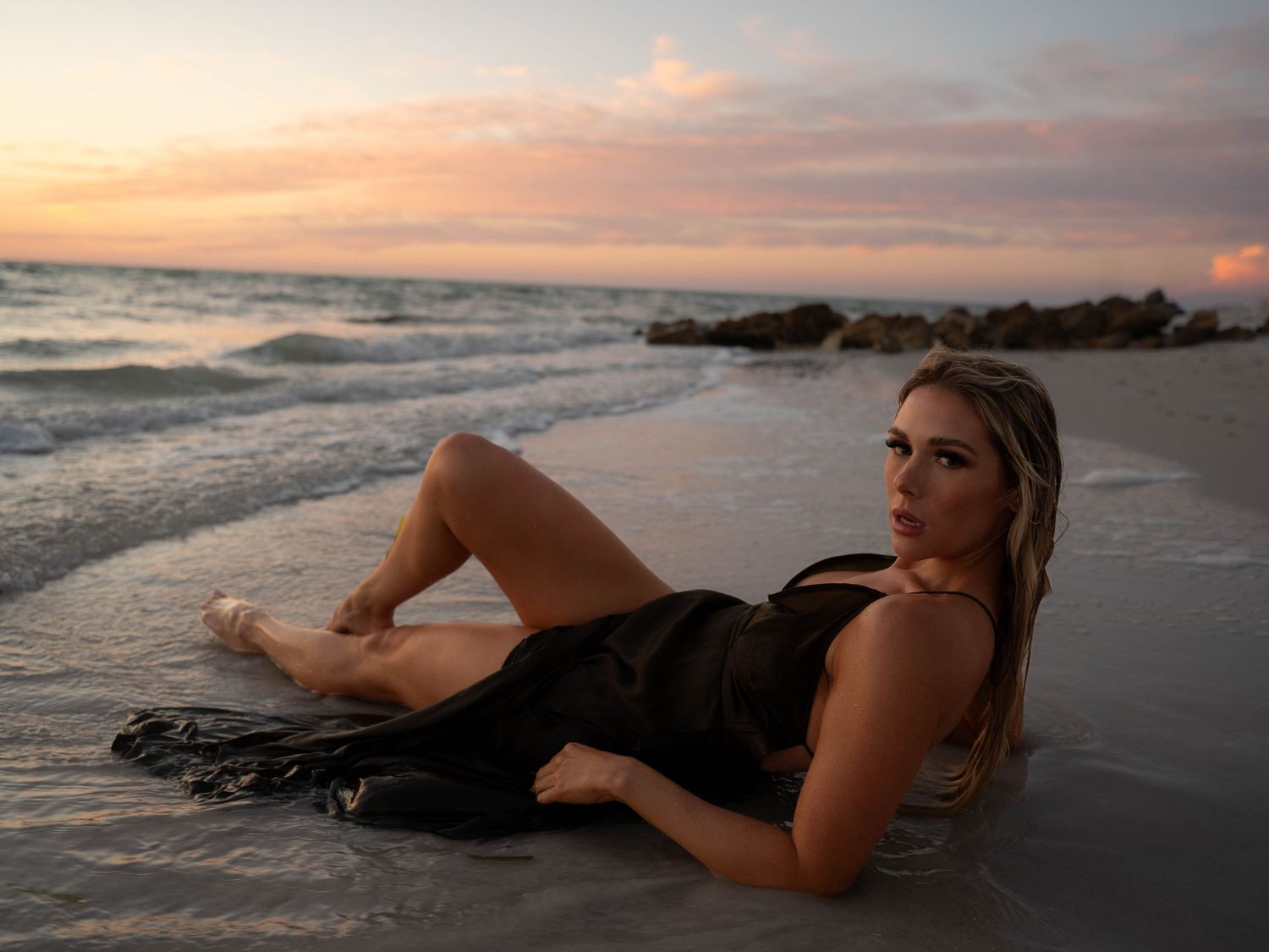 Woman in dark fringed dress poses lying in ocean waves on sandy beach at sunset.