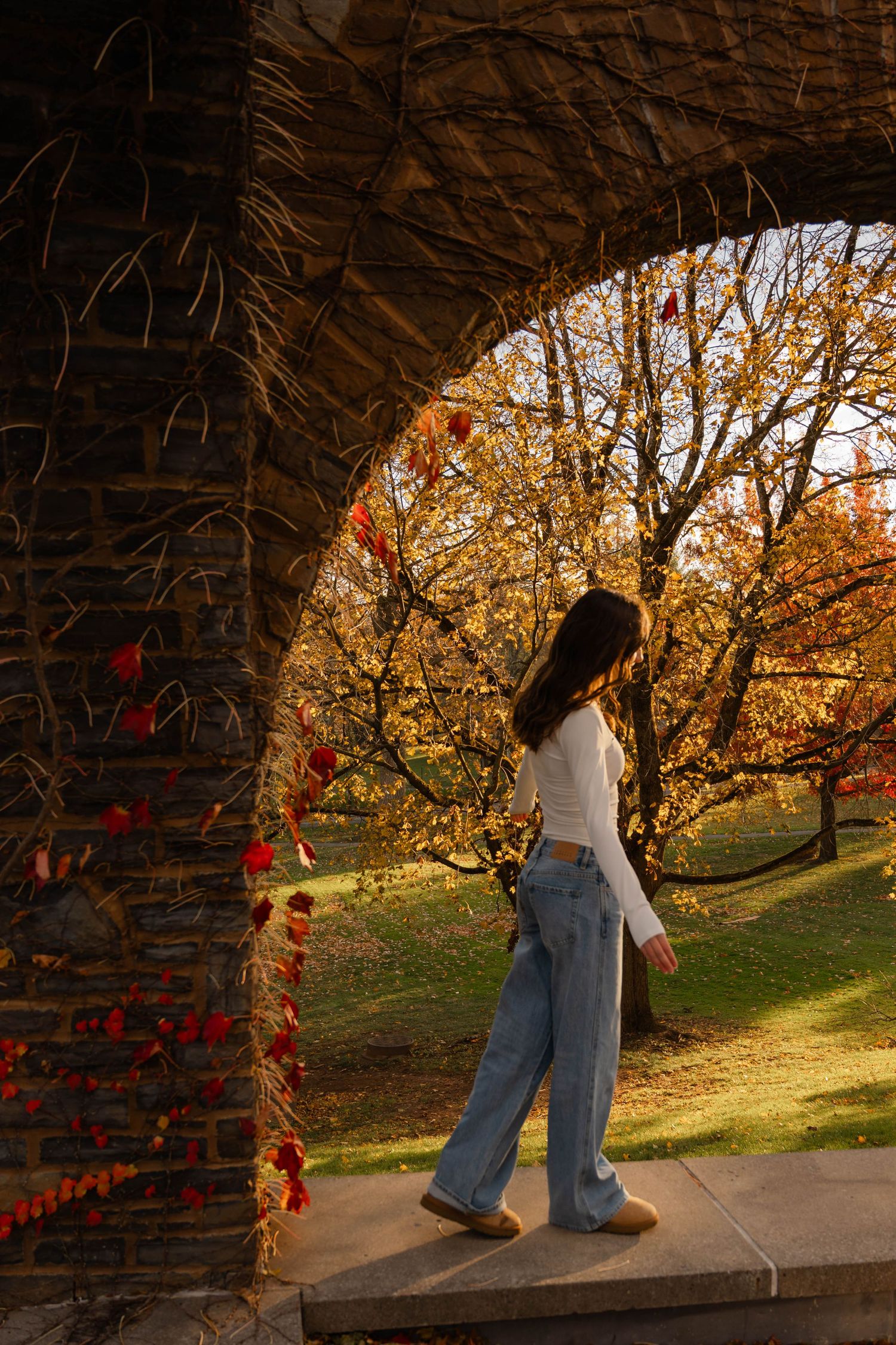 Teen girl in white top and jeans walking under stone arch surrounded by colorful autumn foliage.