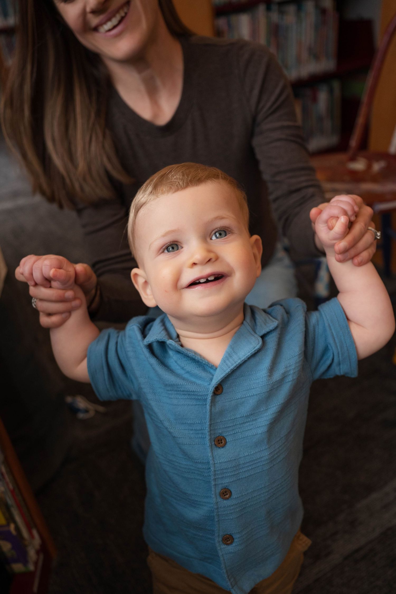 A happy baby boy in a blue shirt stands with arms raised, supported by his mother in a library.