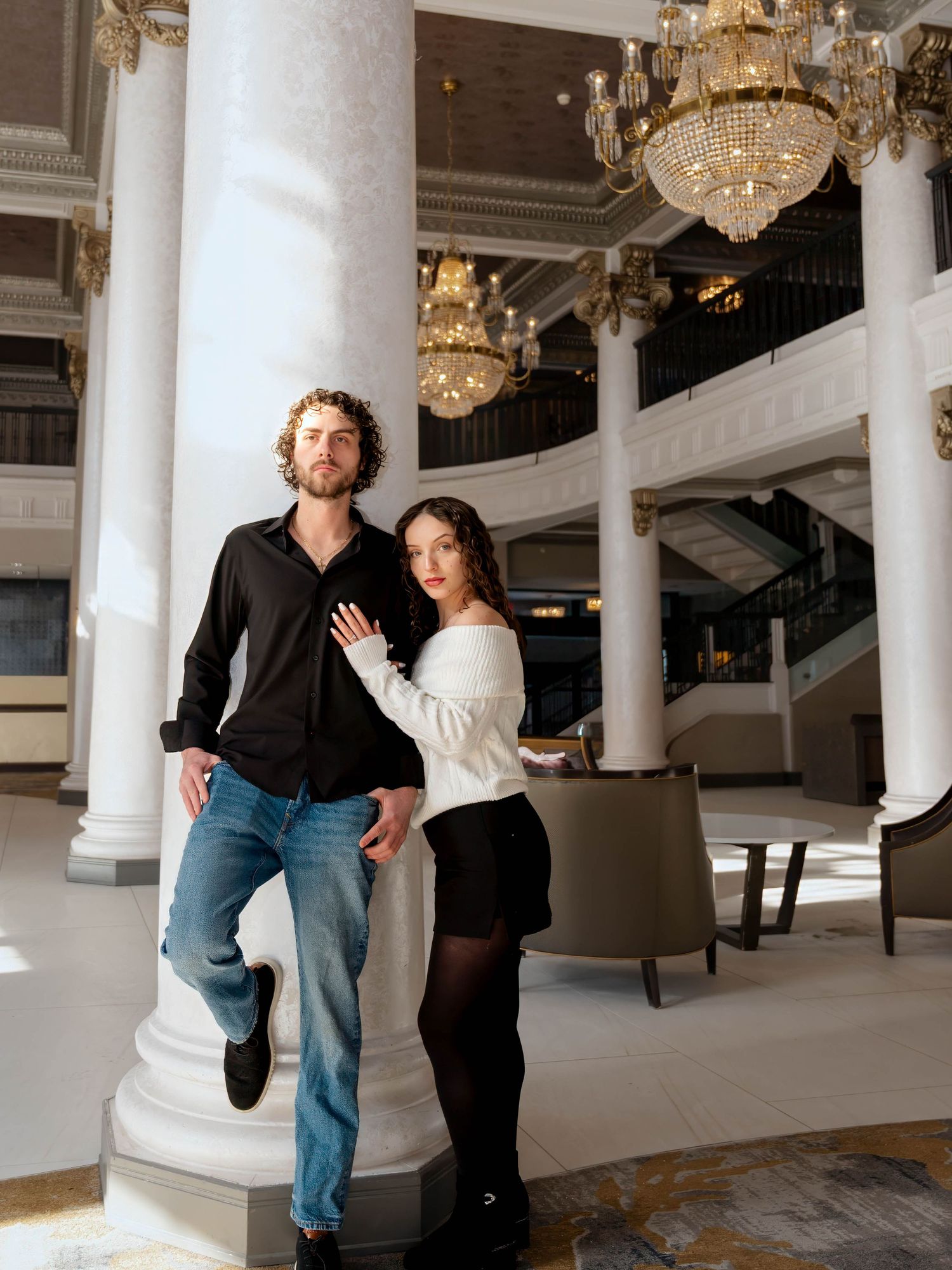 An elegant couple poses together beside a white column in a grand hotel lobby with chandeliers.