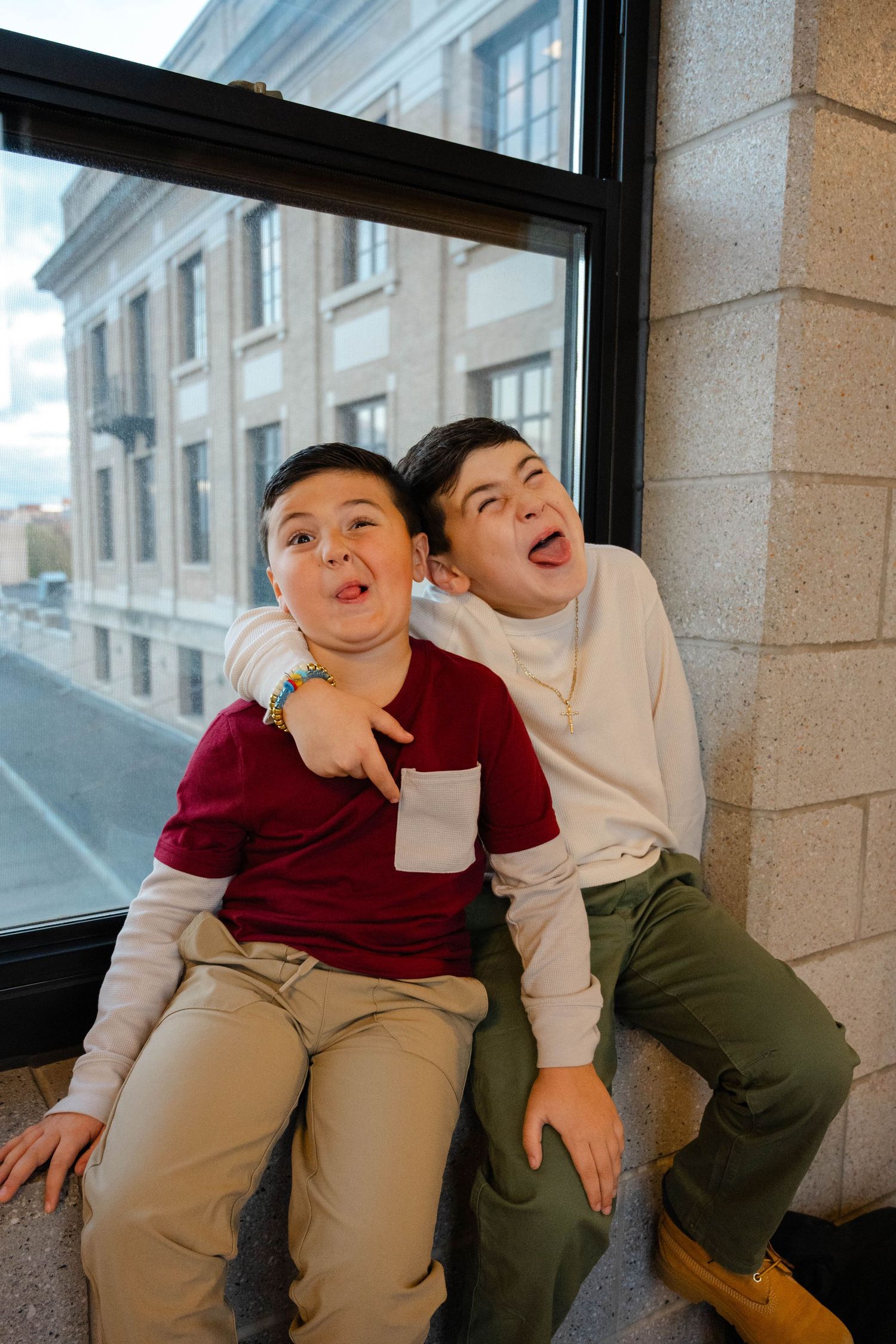 Two playful young brothers make silly faces while sitting on a ledge by a large window outside.