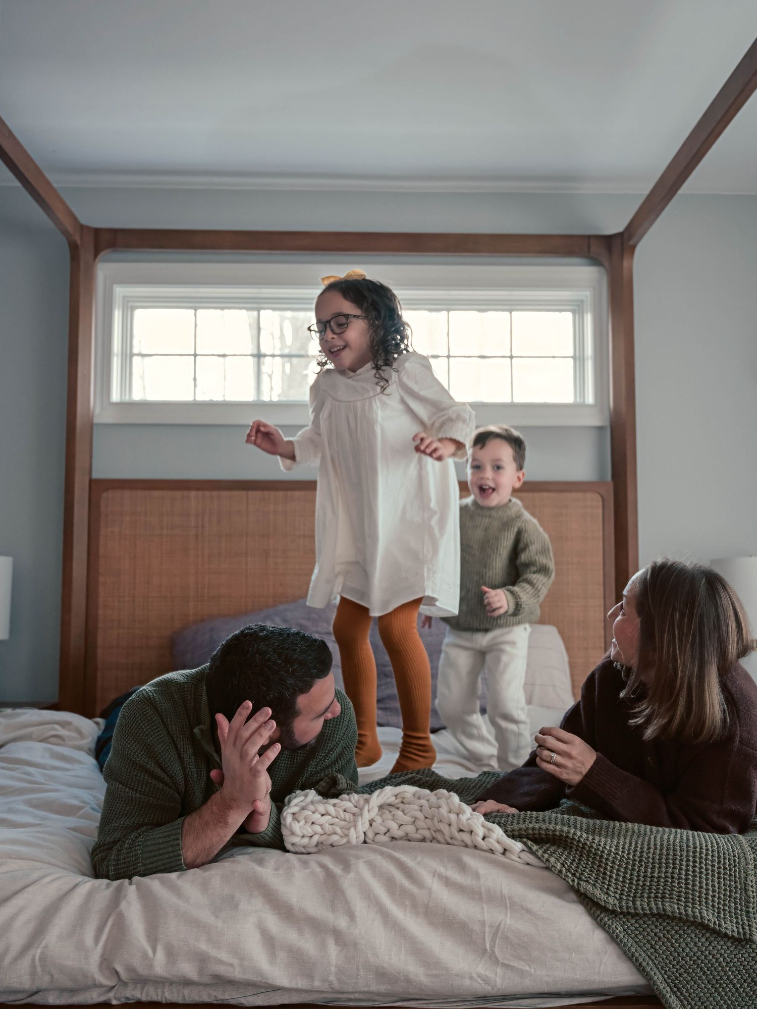Two energetic children jump on a bed while their parents laugh and play along with them.