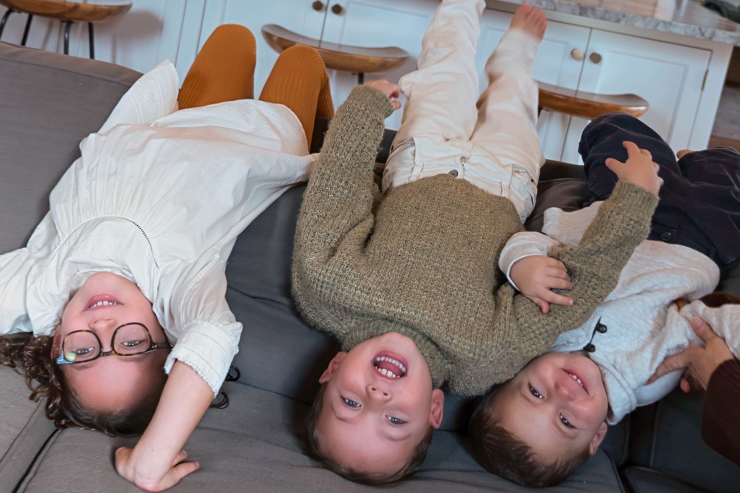 Three children lying on their backs on a couch laughing together in a candid color photo.