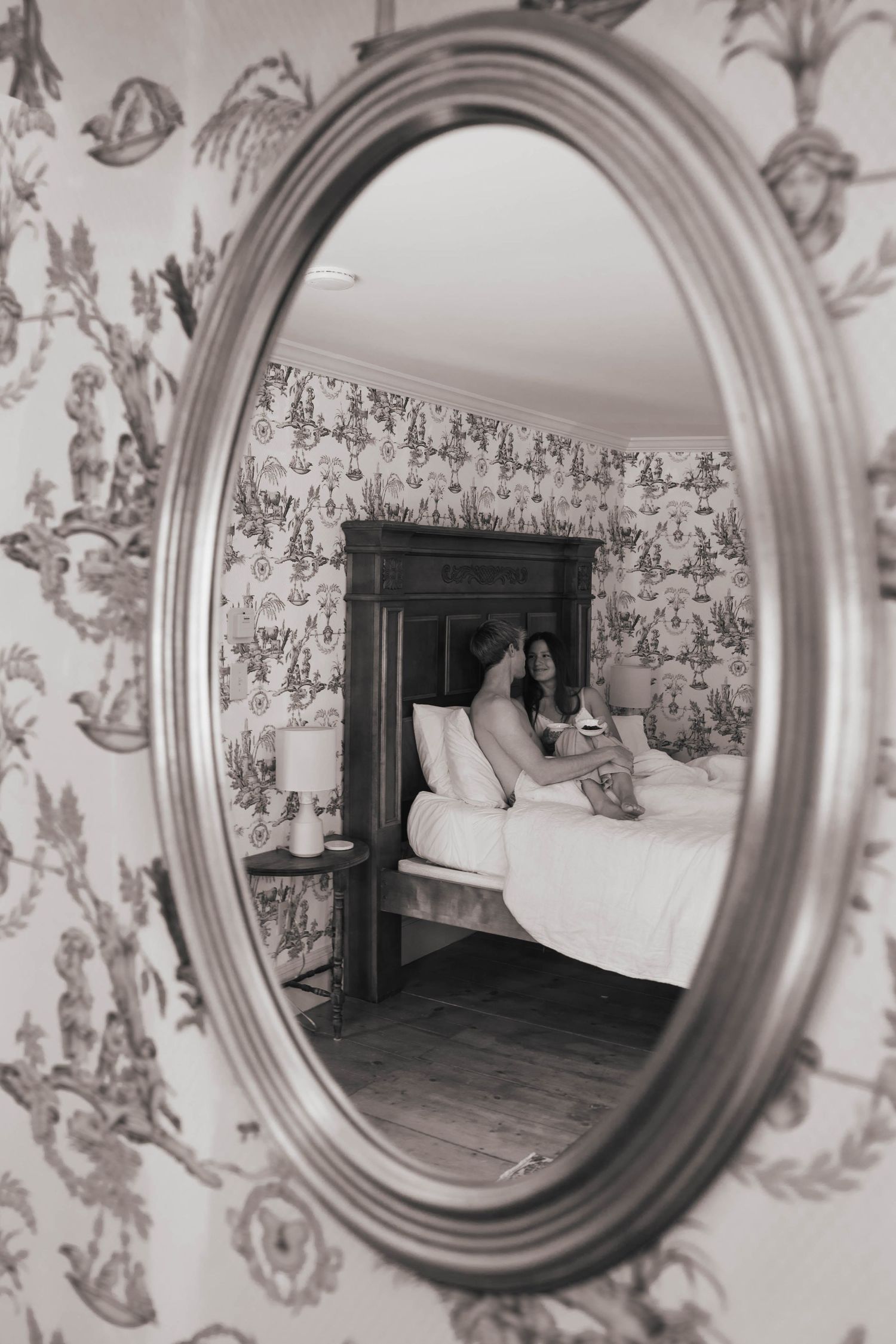 Oval vintage mirror reflecting couple relaxing in bed within elegantly wallpapered bedroom.