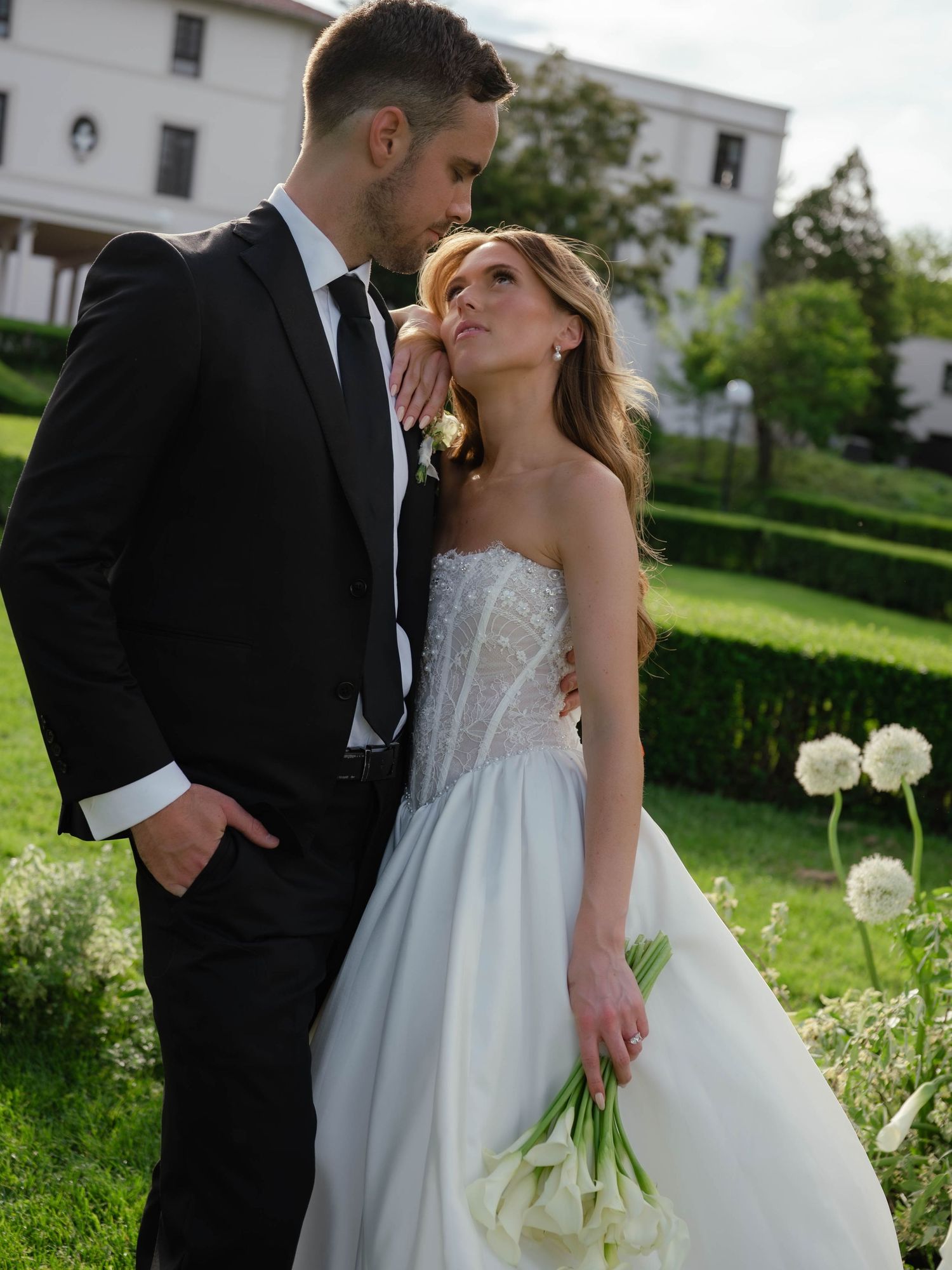 Groom in black suit kisses bride wearing strapless beaded gown holding white calla lilies in lush garden.