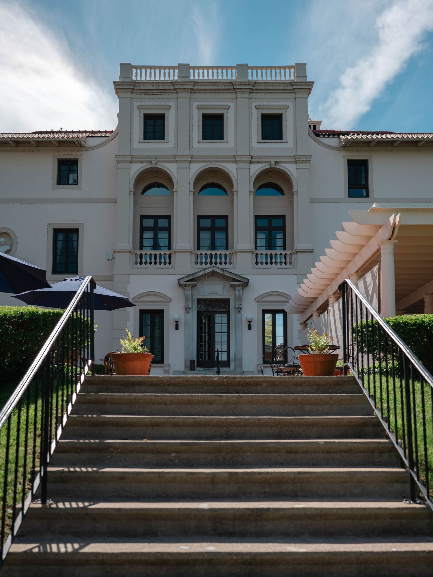 Grand neoclassical mansion exterior with stone staircase, arched windows, and manicured hedges under blue sky.