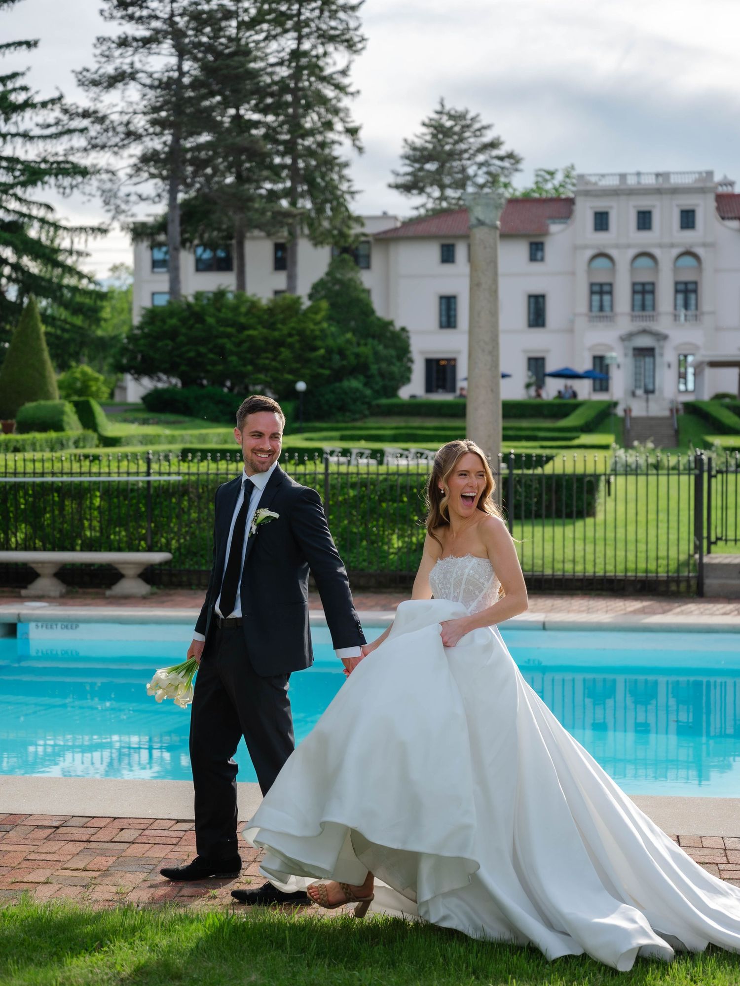 Laughing bride in ballgown and groom in dark suit stroll poolside with grand estate building in background.