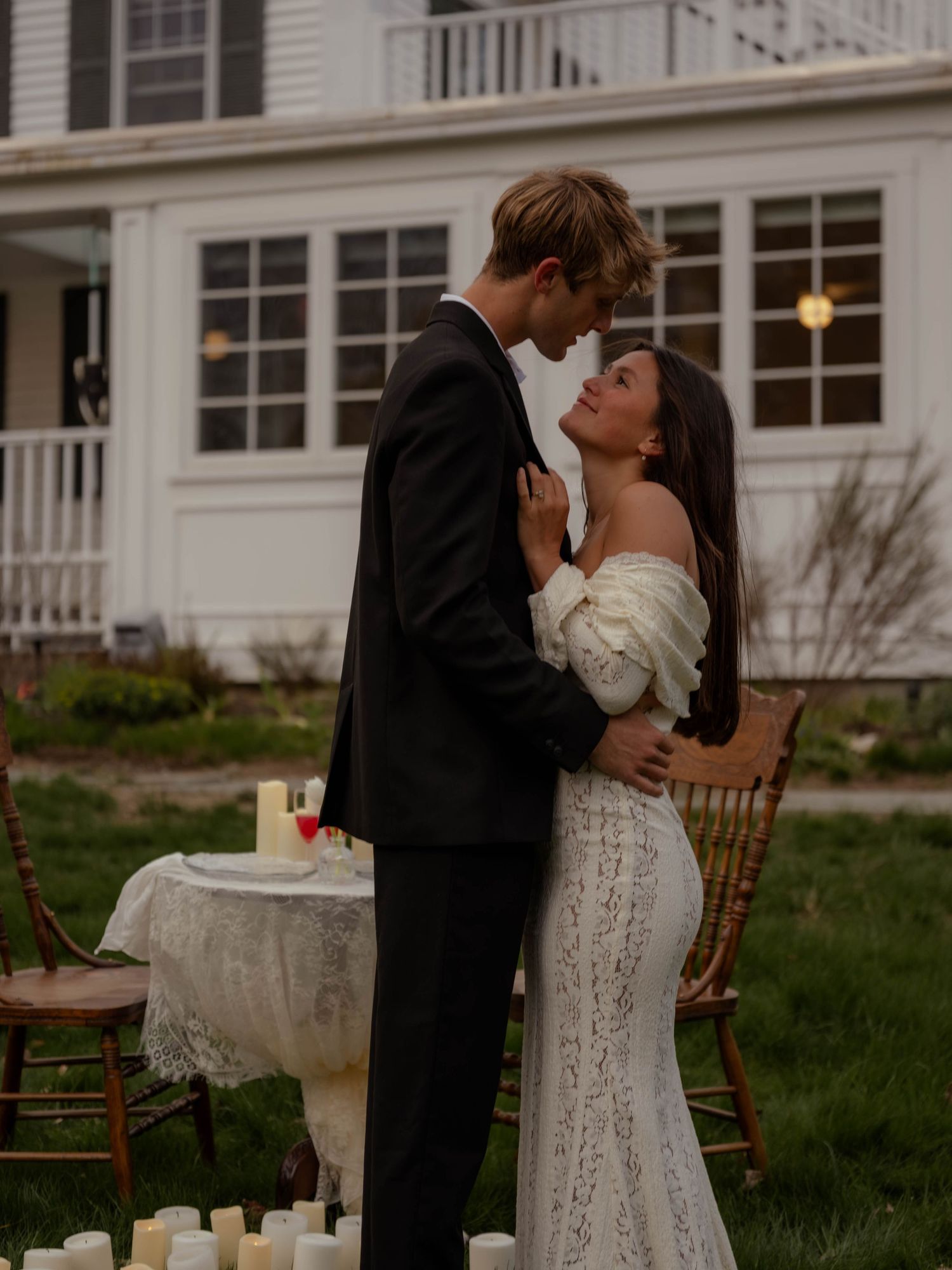 Couple embracing outdoors at dusk near candles, groom in black suit and bride in off-shoulder lace gown.