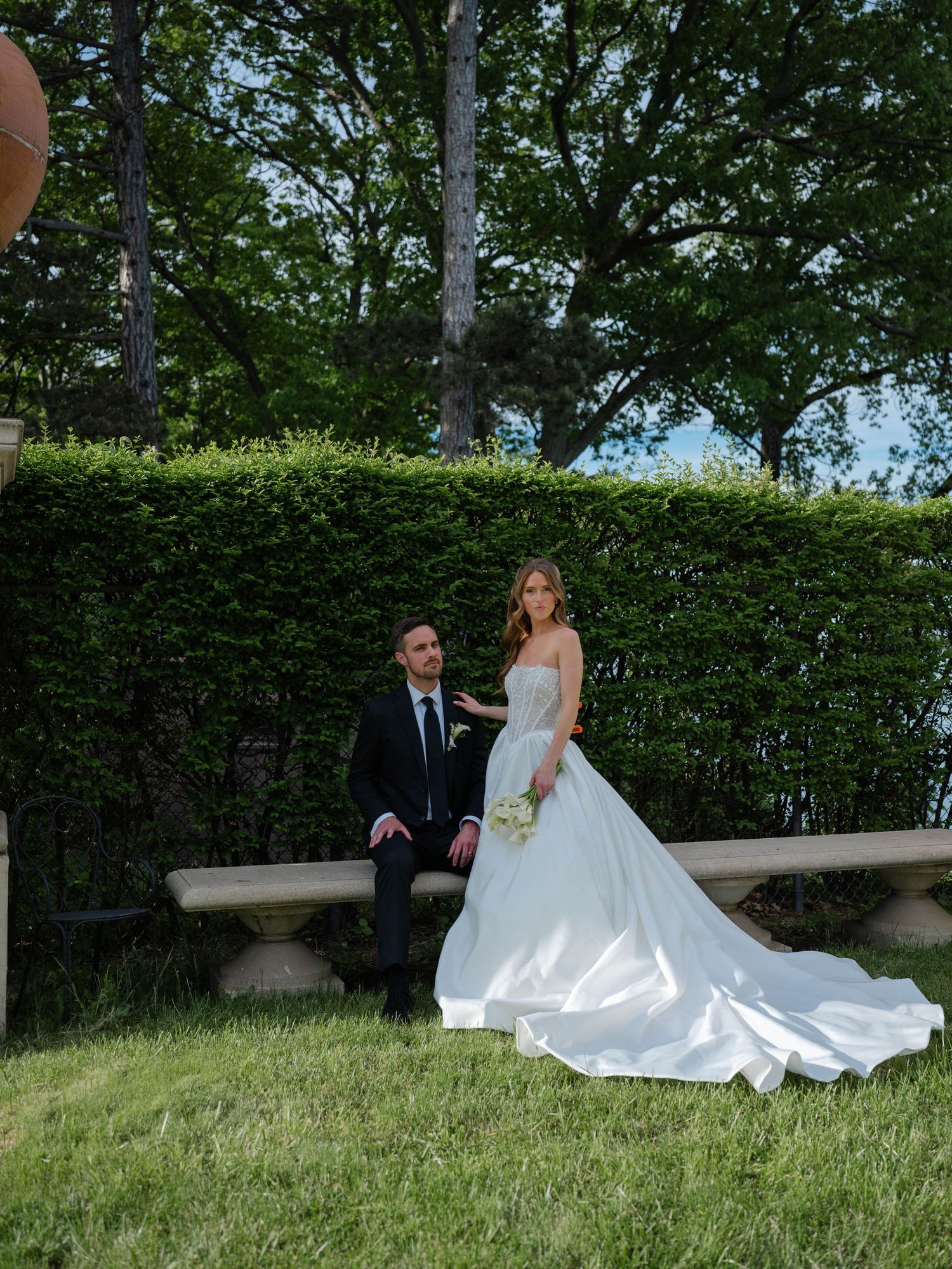 Bride in flowing white gown and groom in black suit pose on a stone bench in a manicured garden.