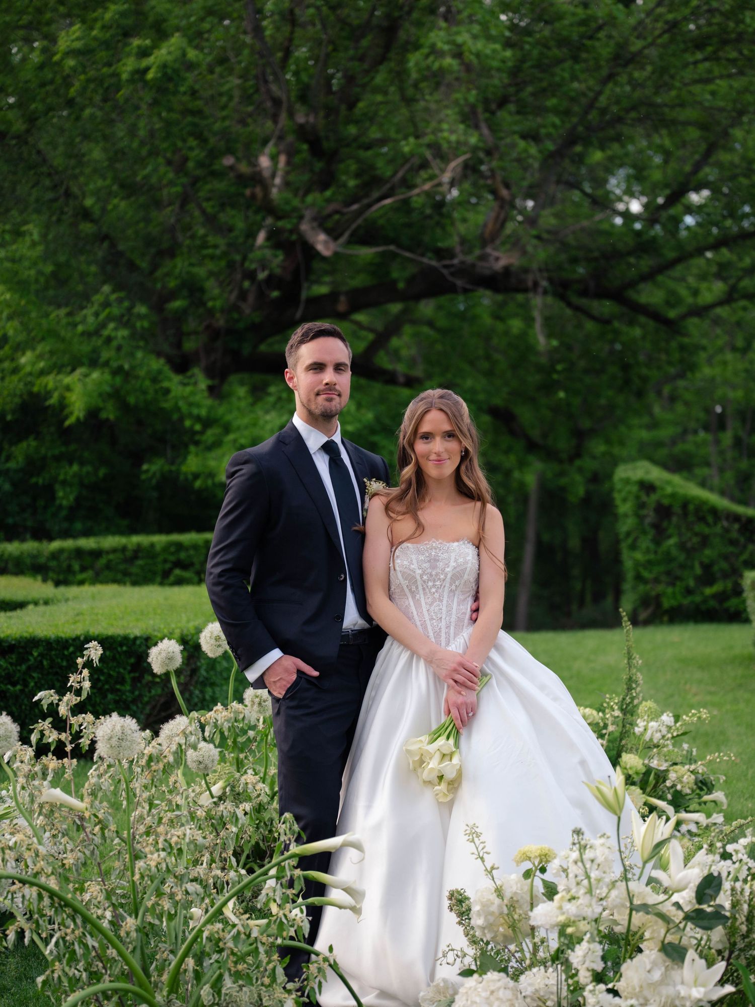 Bride and groom pose together among white blooming flowers with tall trees and green lawn behind them.