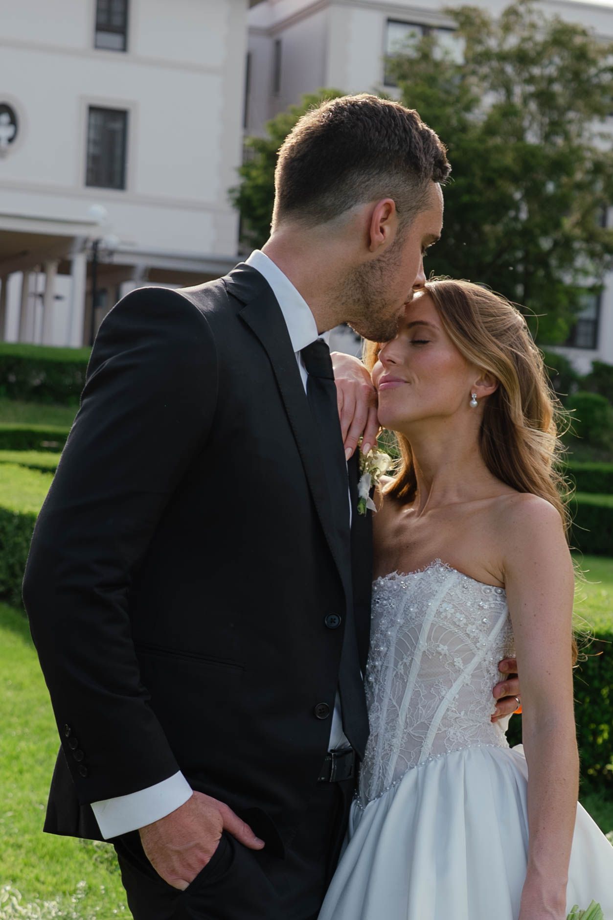 Groom leans in to kiss bride in strapless beaded wedding gown in sunlit garden of elegant manor.